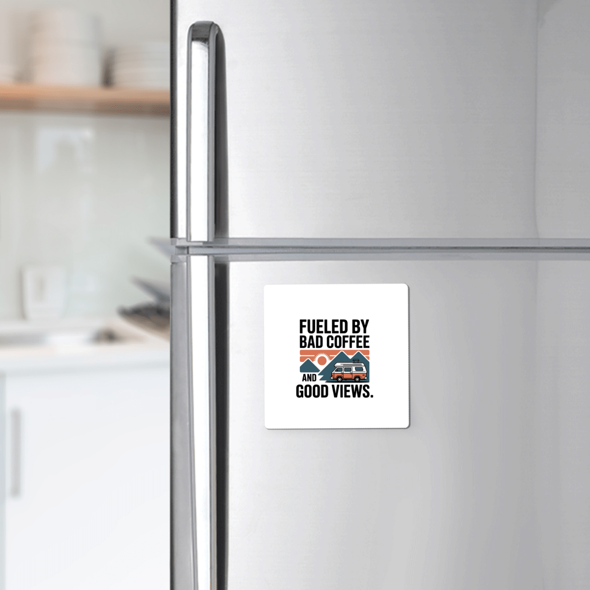 A silver refrigerator displays the "Fueled by Bad Coffee and Good Views Magnet" from Mirabilia Boutique—a fun camping souvenir with a coffee mug and mountain graphic—above a kitchen counter with dishes in the background.
