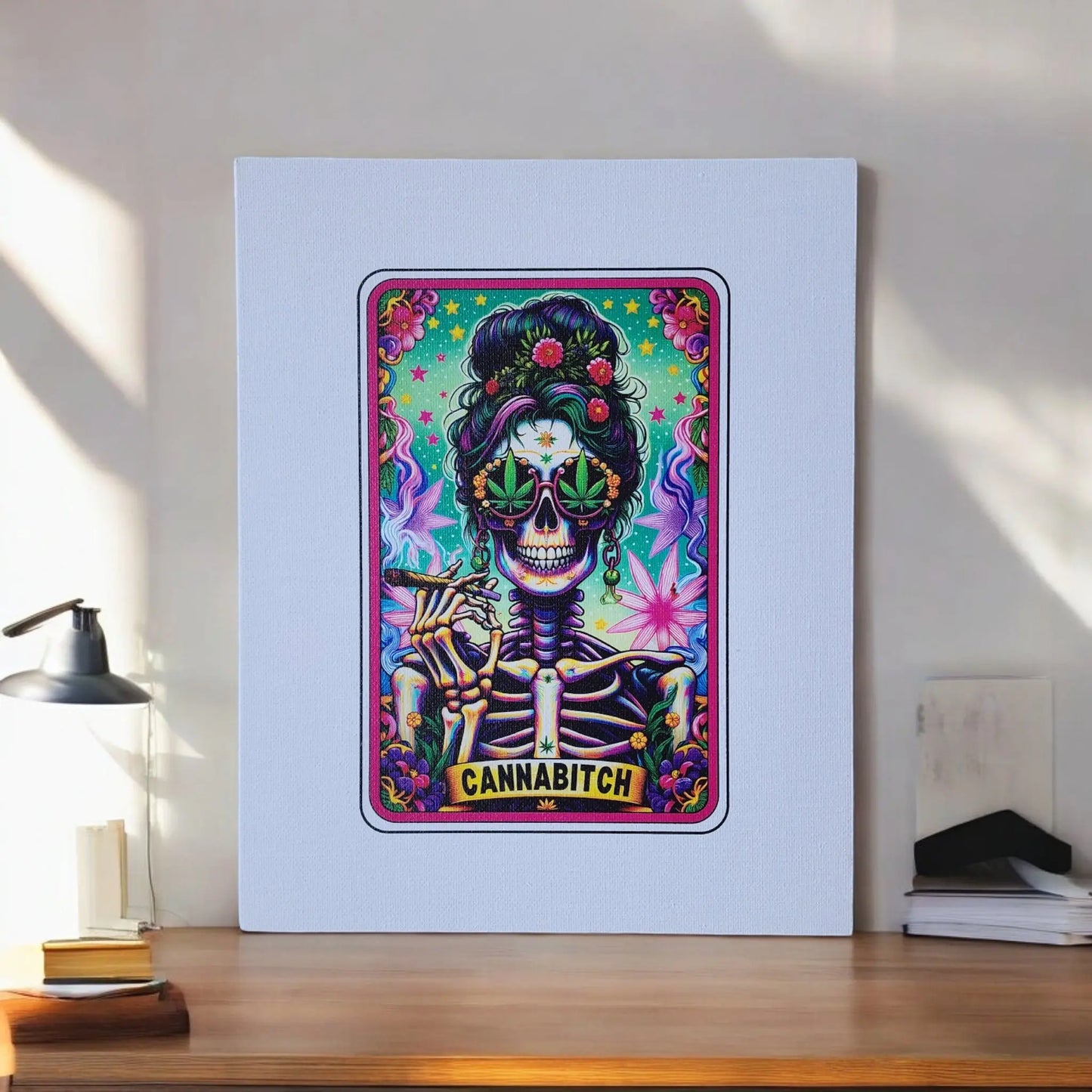 A Cannabitch 8x10 Canvas Sign from Mirabilia Boutique is displayed on a table against a wall. This vibrant artwork features a skeleton adorned with colorful flowers in its hair and sporting heart-shaped glasses. The skeleton is smoking, with the text "CANNABITCH" written at the bottom, all set against a bright, psychedelic background.