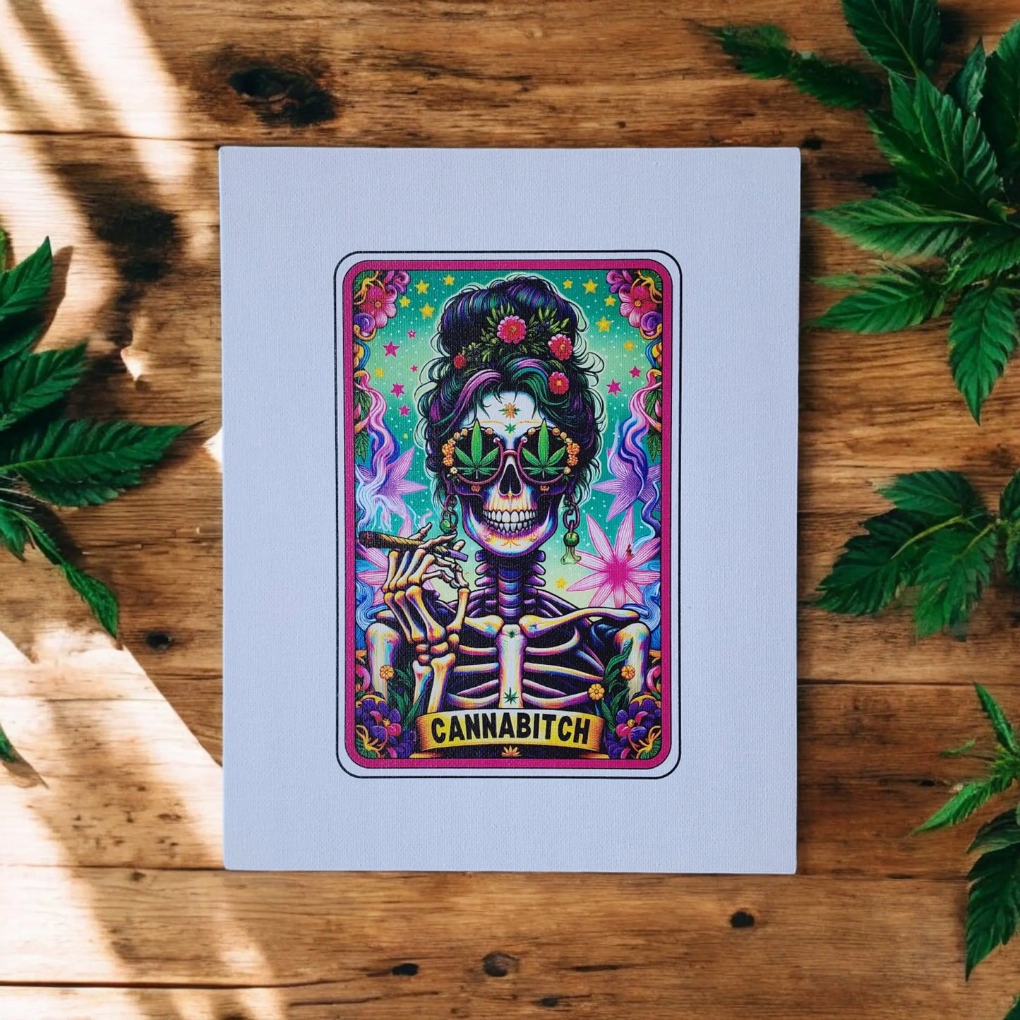 A vibrant, psychedelic painting on a white canvas depicts a skeleton wearing sunglasses and adorned with flowers and leaves. Below the skeleton is the word "CANNABITCH." This Cannabitch 8x10 Canvas Sign from Mirabilia Boutique, surrounded by wooden surfaces and green leaves, embodies cannabis culture art with a tarot card design flair.