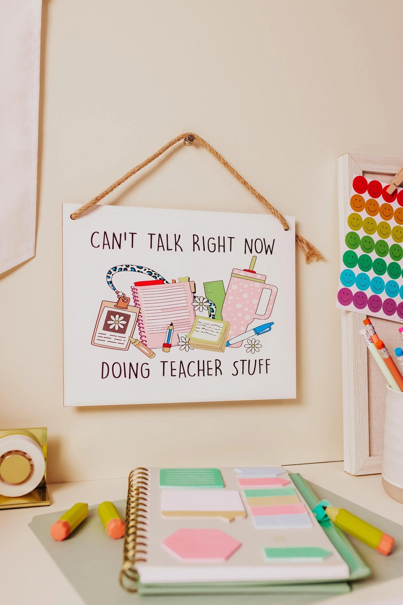 In a classroom setting adorned with vibrant decor, a sign from Mirabilia Boutique titled "Can't Talk Right Now: Teacher Stuff" hangs prominently. Surrounding it, you'll find colorful pens, a notebook filled with sticky notes, and a wooden shelf decorated with smiley magnets—perfect additions for any teaching environment.