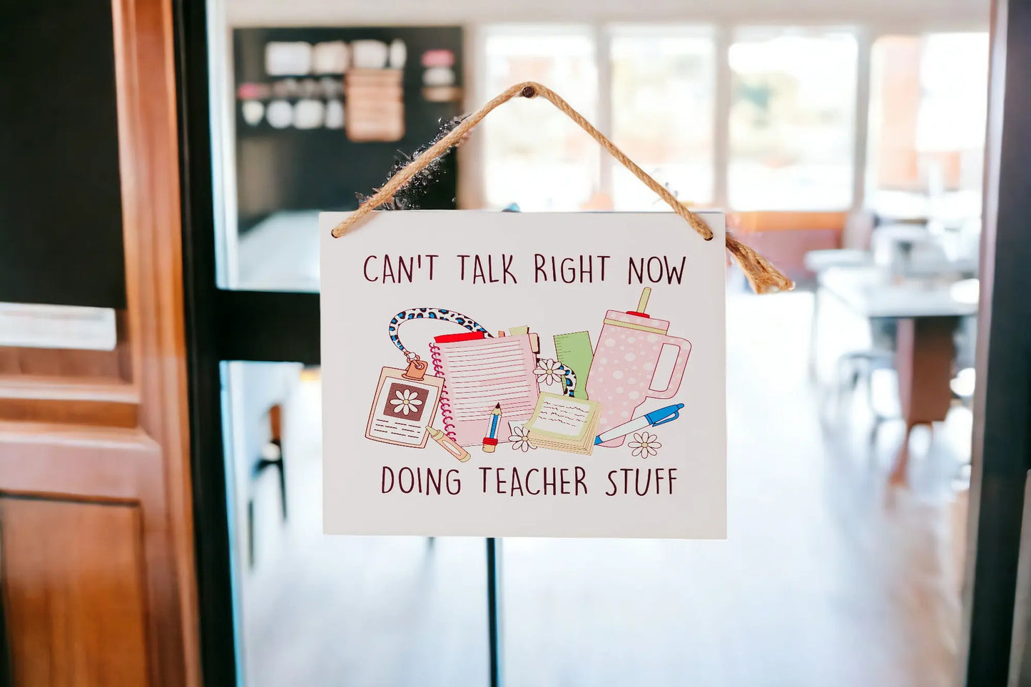 A whimsical touch is added to the classroom with Mirabilia Boutique's "Can't Talk Right Now: Teacher Stuff Hanging Sign." Adorned with illustrations of a coffee cup, planner, and pencils, this delightful piece complements the softly blurred background of a bustling classroom.