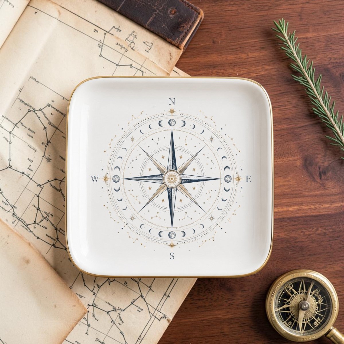 The Celestial Compass Jewelry Tray | Gilded Ceramic by Mirabilia Boutique, adorned with a compass rose and moon phases design, rests on a wooden table alongside a map, compass, leather-bound book, and a sprig of greenery—an elegant way to infuse your space with celestial decor.