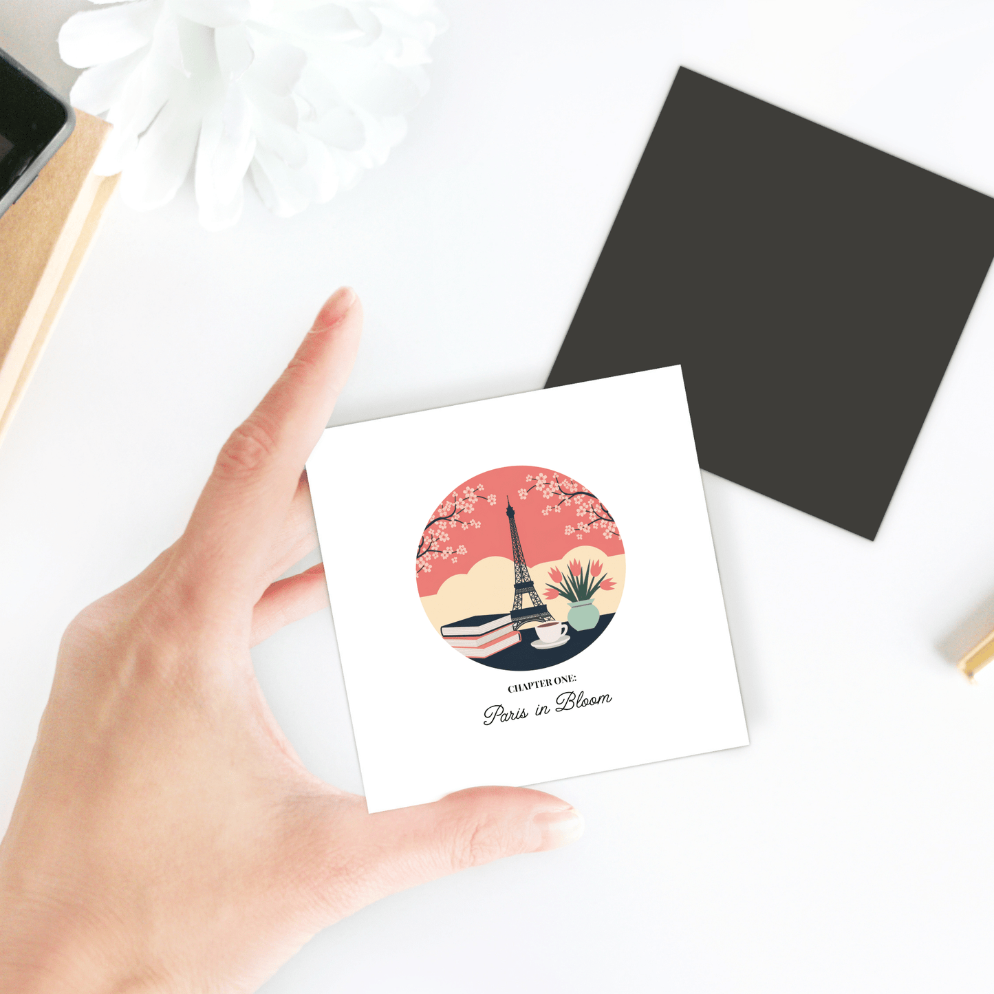 A hand holds the Paris in Bloom Magnet by Mirabilia Boutique, featuring a floral Eiffel Tower illustration and “CAPTURED: Paris in Bloom”—a perfect literary Paris gift for book lovers. A black envelope and white flower sit nearby.