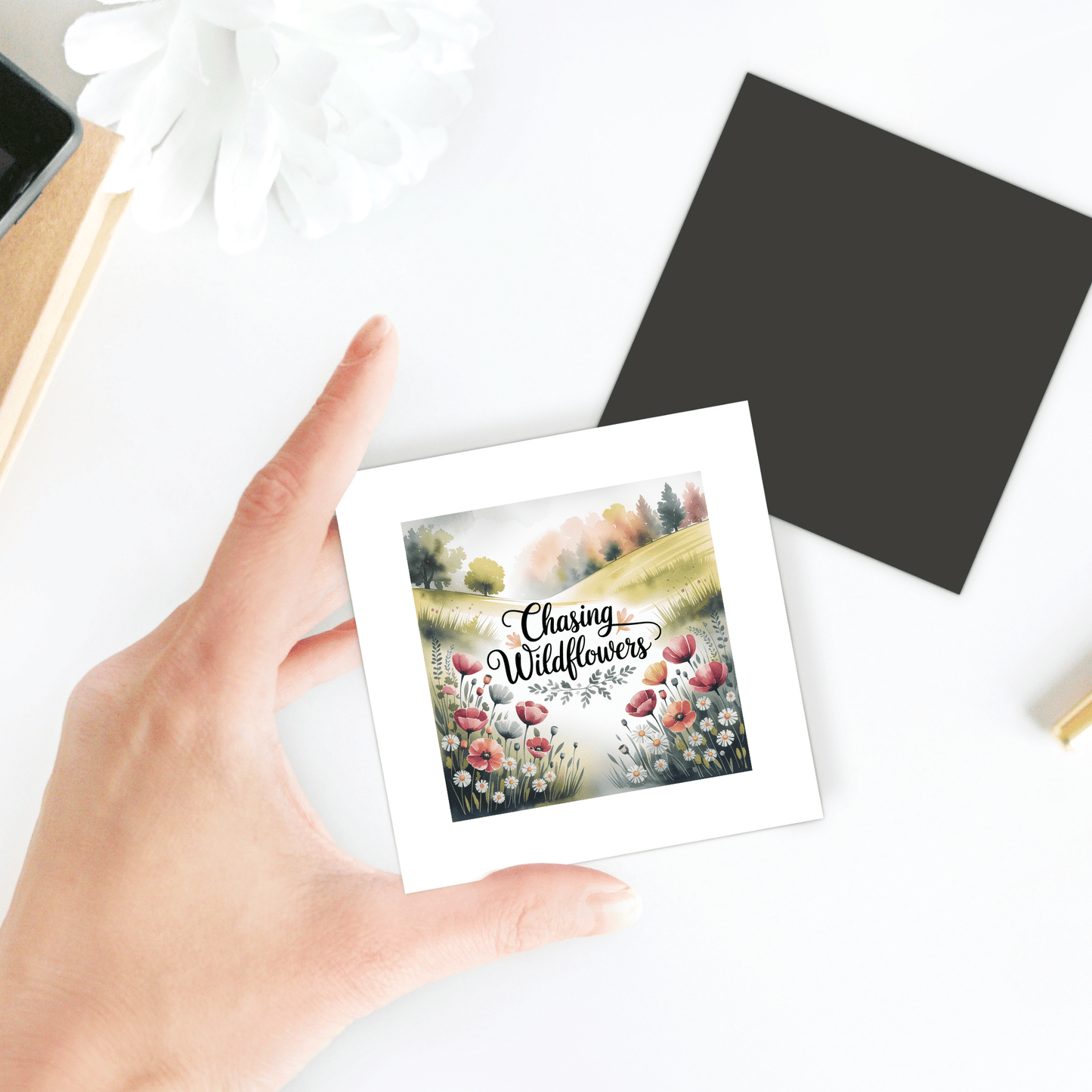 A hand holds the Chasing Wildflowers Magnet by Mirabilia Boutique, featuring a colorful wildflower field and nature quote. Other stationery is visible on a white desk in the background.