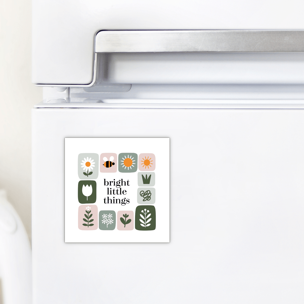 A white refrigerator is decorated with the Mirabilia Boutique Bright Little Things Floral Grid Magnet, featuring simple floral and nature illustrations, a bee, and the words "bright little things" in the center.