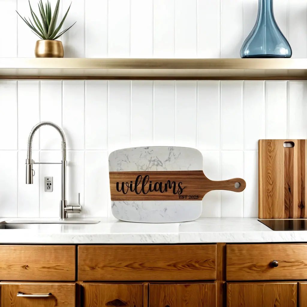 A modern kitchen features wood cabinets, a Mirabilia Boutique Personalized Marble and Acacia Serving Board engraved with “williams EST 2023,” a plant in a gold pot, tall blue vase, silver faucet, and white tile backsplash.