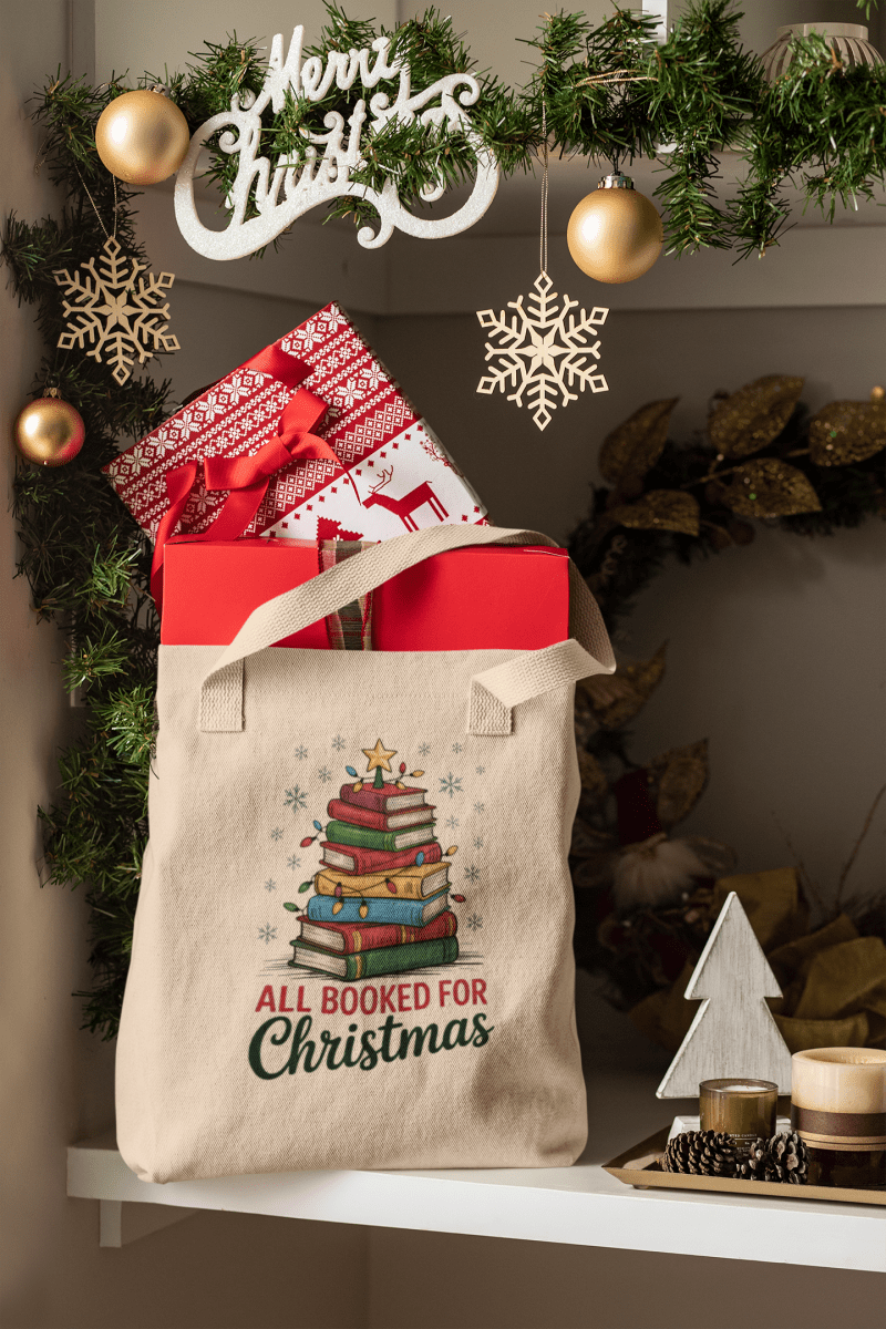 A festive scene with the Mirabilia Boutique "All Booked for Christmas Book Stack Tote Bag," perfect for book lovers, holding wrapped gifts among ornaments and a "Merry Christmas" sign.