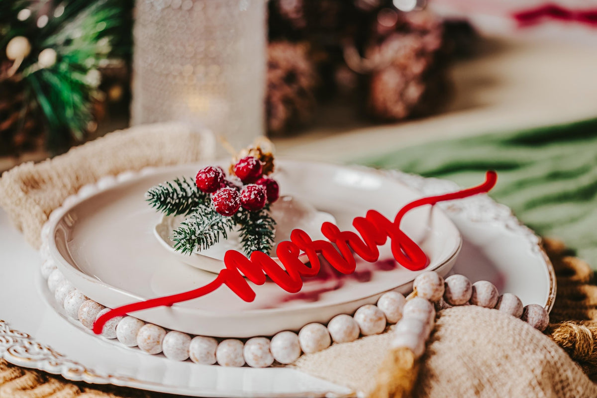 Create a festive holiday table with white plates, beaded garland, pine and berry decor, and Mirabilia Boutique’s Red Acrylic Christmas Plate Words—an 8-piece set that adds charming script to each plate.