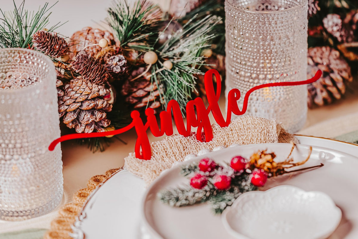A festive holiday table features pinecones, greenery, and candle holders, with the Mirabilia Boutique Red Acrylic Christmas Plate Words from the 8 Piece Holiday Table Decor Set as a jolly “jingle” accent atop each white plate.