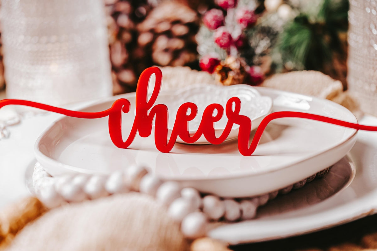 The Mirabilia Boutique Red Acrylic Christmas Plate Words 8-piece set features the decorative word "cheer" in red script, displayed on a white plate among festive table settings with blurred ornaments and greenery.