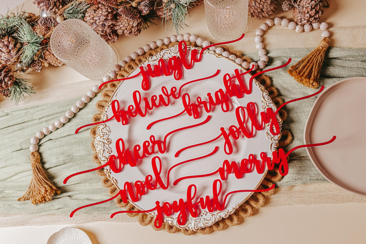 A holiday table shines with Mirabilia Boutique’s Red Acrylic Christmas Plate Words—an 8-piece set featuring festive script like "jingle," "believe," and "cheer"—surrounded by pinecones, beads, and glasses for a bright Christmas display.