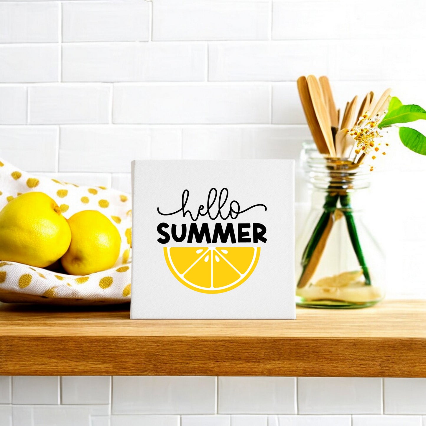 The Mirabilia Boutique Hello Summer Lemon Canvas Sign – 8x8 Wall Art brightens a wooden shelf with cheerful lemon decor—a bowl of lemons and a jar with utensils and flowers—adding fresh summer vibes to a white tiled wall.