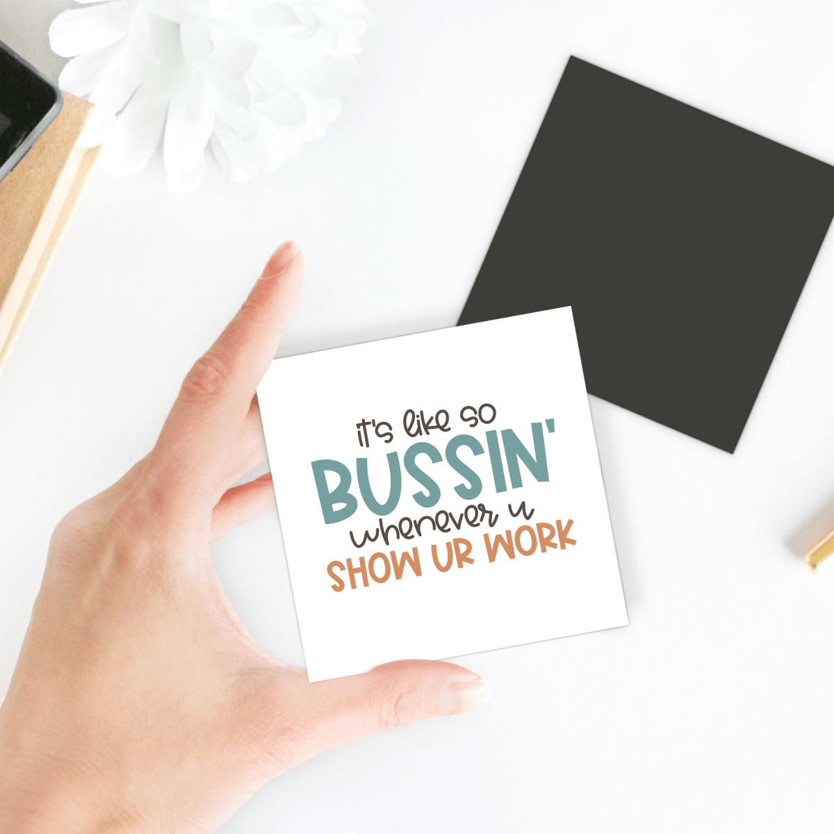 A hand holds Mirabilia Boutique's "It's So Bussin' Show Your Work Magnet," perfect for classroom decor or as a fun teacher gift. In the background: a white flower, black square, and notepad.