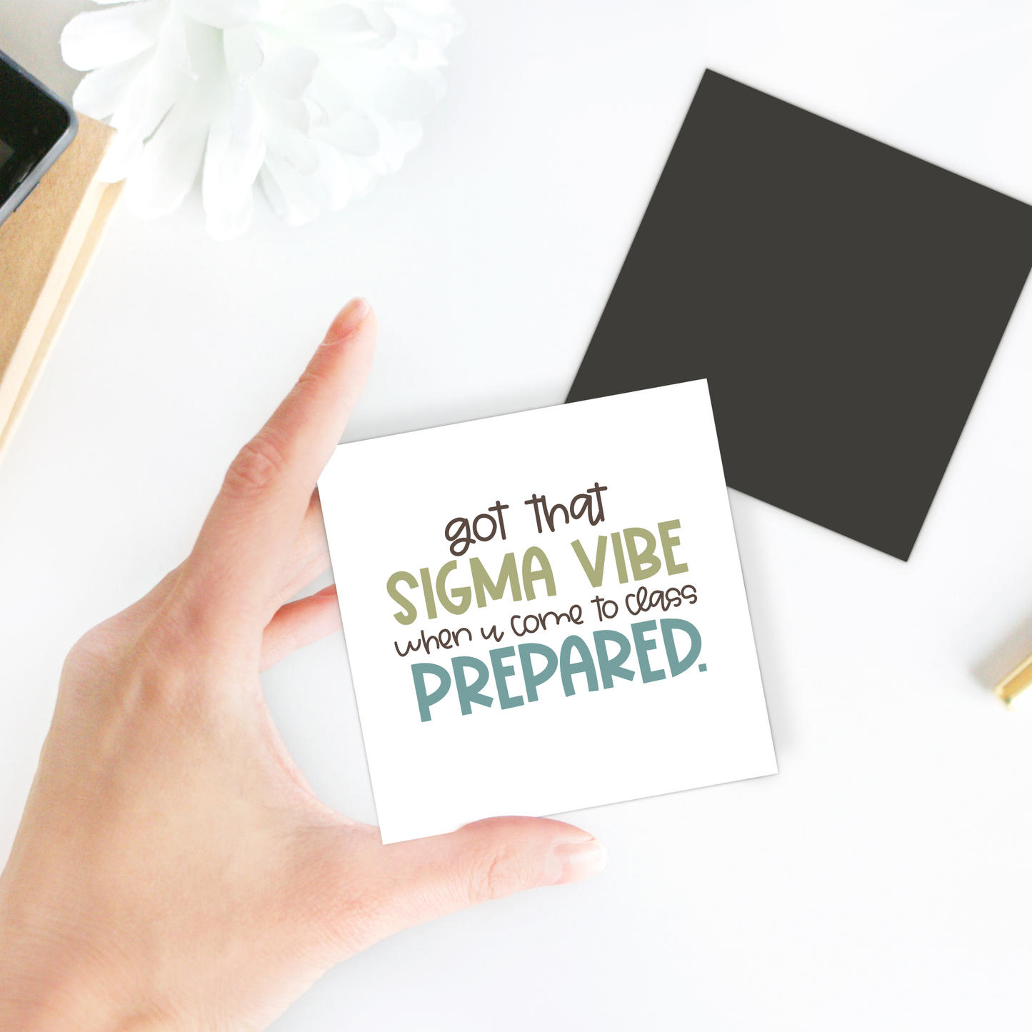 A hand holds the "Got That Sigma Vibe Classroom Magnet" by Mirabilia Boutique, featuring the quote "Got that sigma vibe when u come to class prepared." Displayed on a white surface with a black square, pen, and white flower.