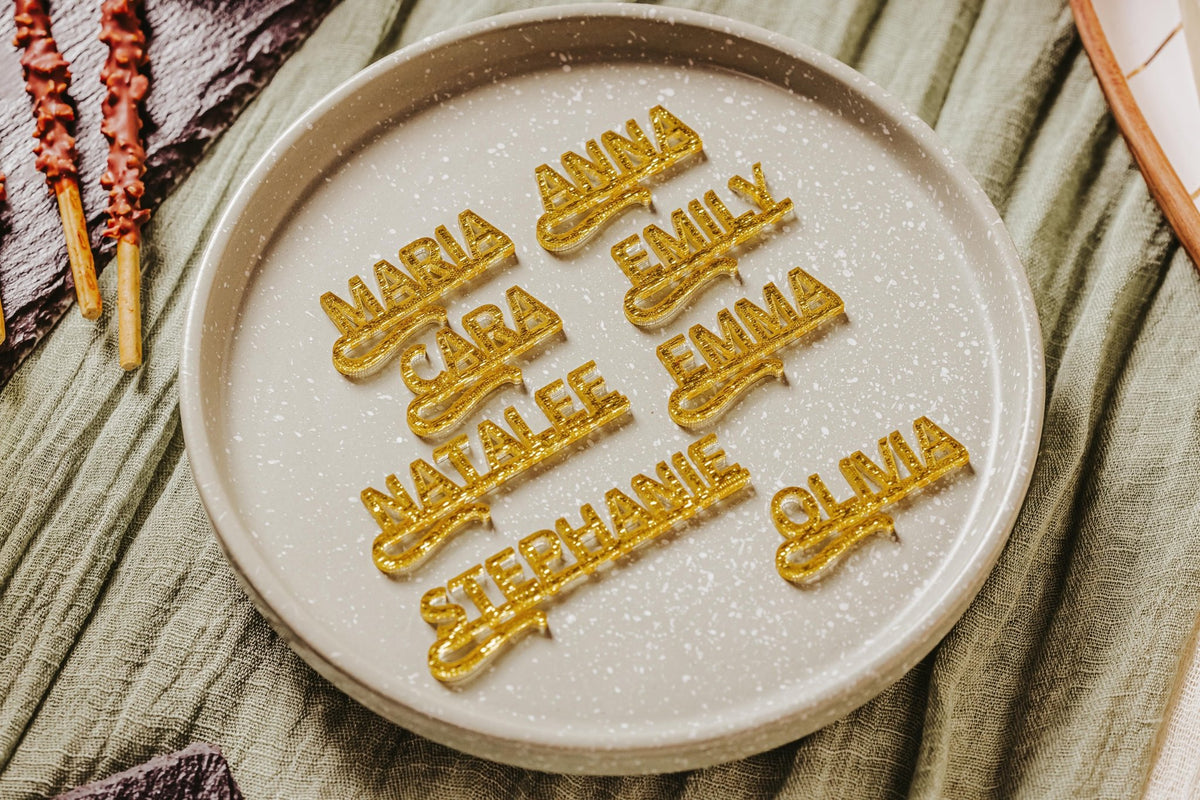 A round beige plate displays eight golden glittery Personalized Wine Glass Name Charms by Mirabilia Boutique—custom acrylic drink tags ideal for bachelorette parties, weddings, or bridal showers—arranged on a speckled surface atop a green cloth.