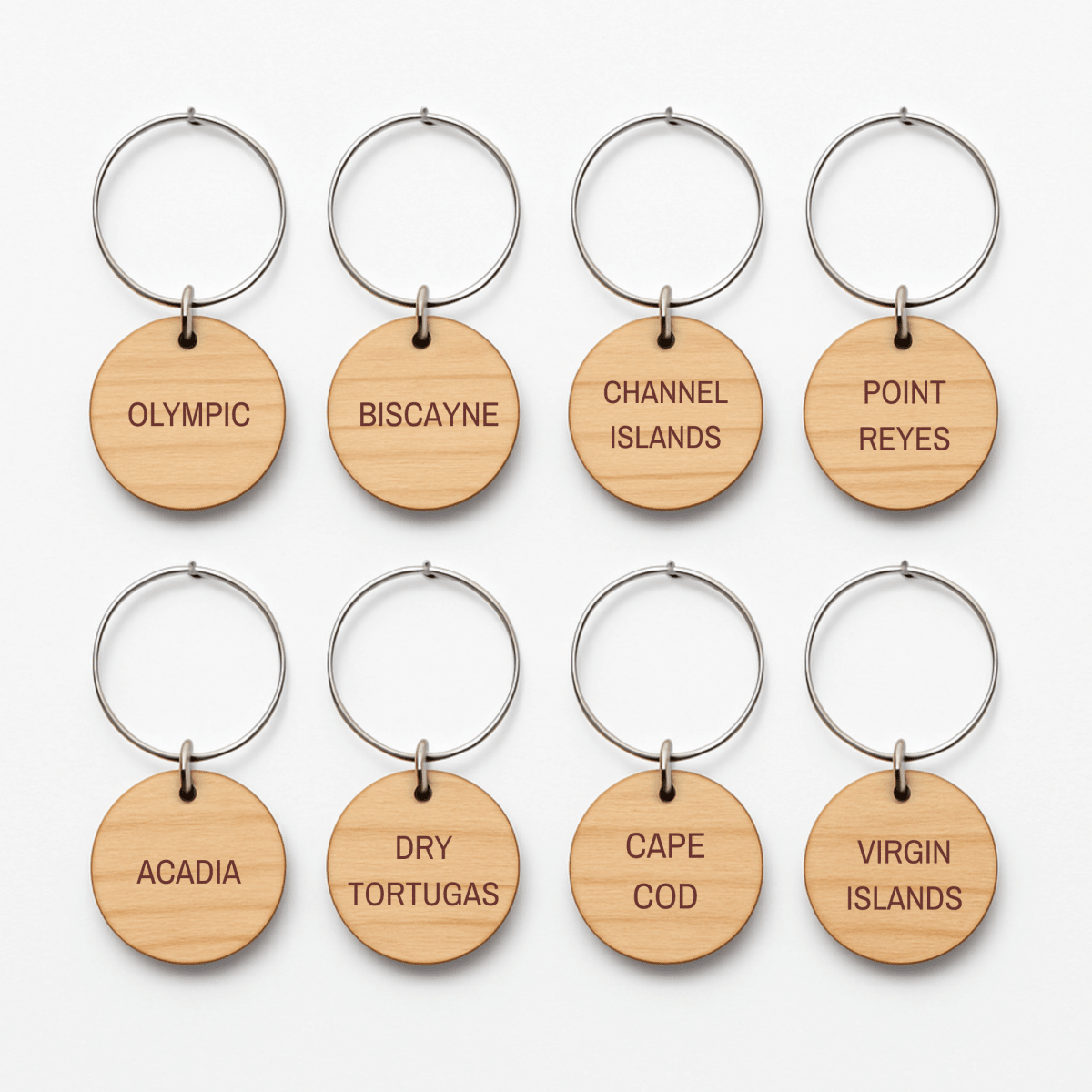 Eight round wooden wine charms with metal rings from Mirabilia Boutique’s Coastal Escapes Collection are arranged in two rows. Each charm is engraved with names of national parks like Olympic, Biscayne, and Channel Islands.