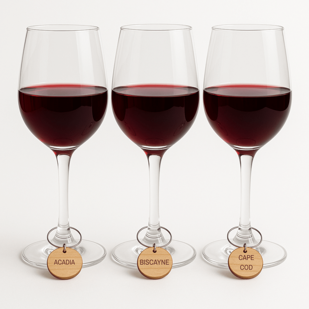 Three red wine glasses with stem tags labeled "ACADIA," "BISCAYNE," and "CAPE COD" display Mirabilia Boutique’s Coastal Escapes Rustic Wine Charms on a white surface.