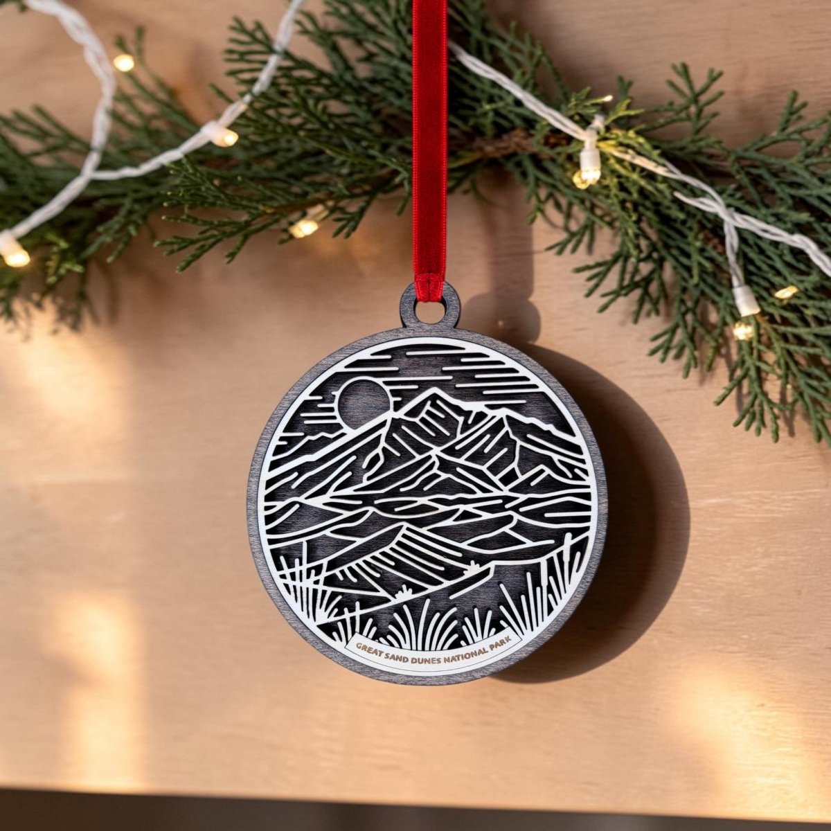 The Great Sand Dunes National Park Line Art Ornament by Mirabilia Boutique is an oval wooden ornament featuring a mountain scene. It hangs from a red ribbon on a garland adorned with greenery and string lights, and is inscribed at the bottom with “Great Sand Dunes National Park.”.