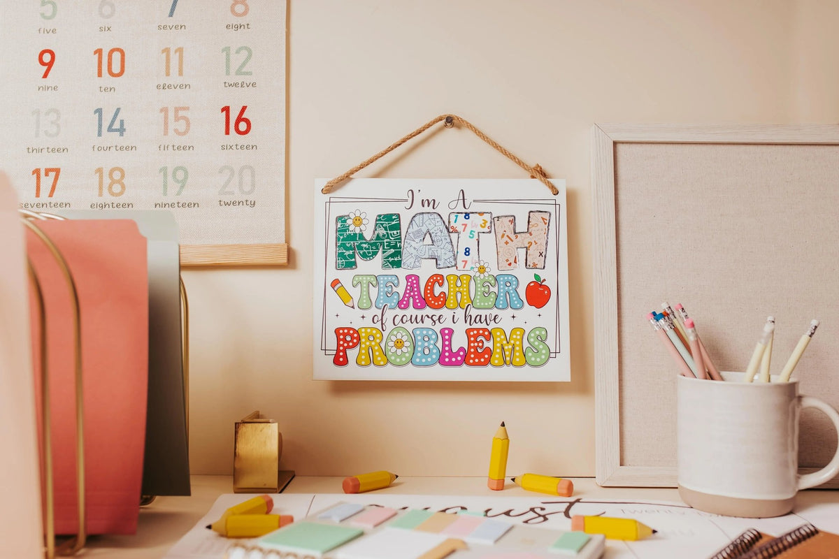 A desk with stationery, colored pencils, erasers, and the Mirabilia Boutique Funny Math Teacher Classroom Sign: "I'm A Math Teacher Of Course I Have Problems," plus math-themed wall decor and scattered papers.