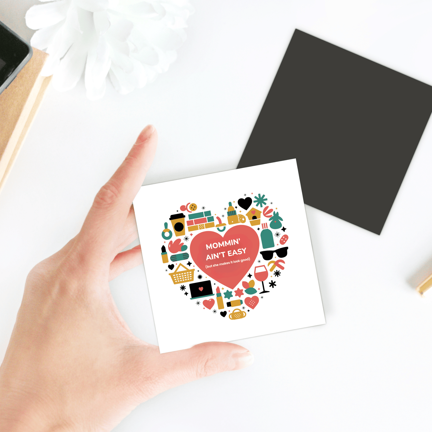 A hand holds the Mommin’ Ain’t Easy Magnet by Mirabilia Boutique, featuring a colorful heart-shaped design—an ideal Mother’s Day gift or cute mom magnet. A black square and white flower rest on the table behind.