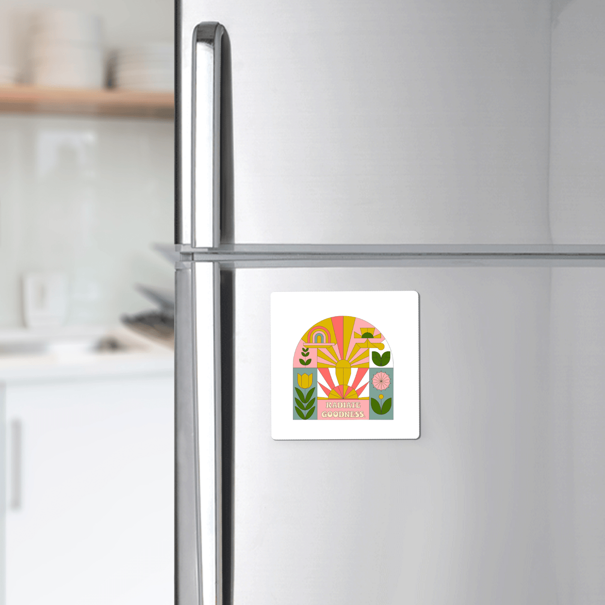 A silver refrigerator features Mirabilia Boutique's Radiate Goodness Colorful Floral Magnet showcasing abstract shapes, leaves, flowers, and the words "RADIATE GOODNESS," set against a bright, tidy kitchen backdrop.