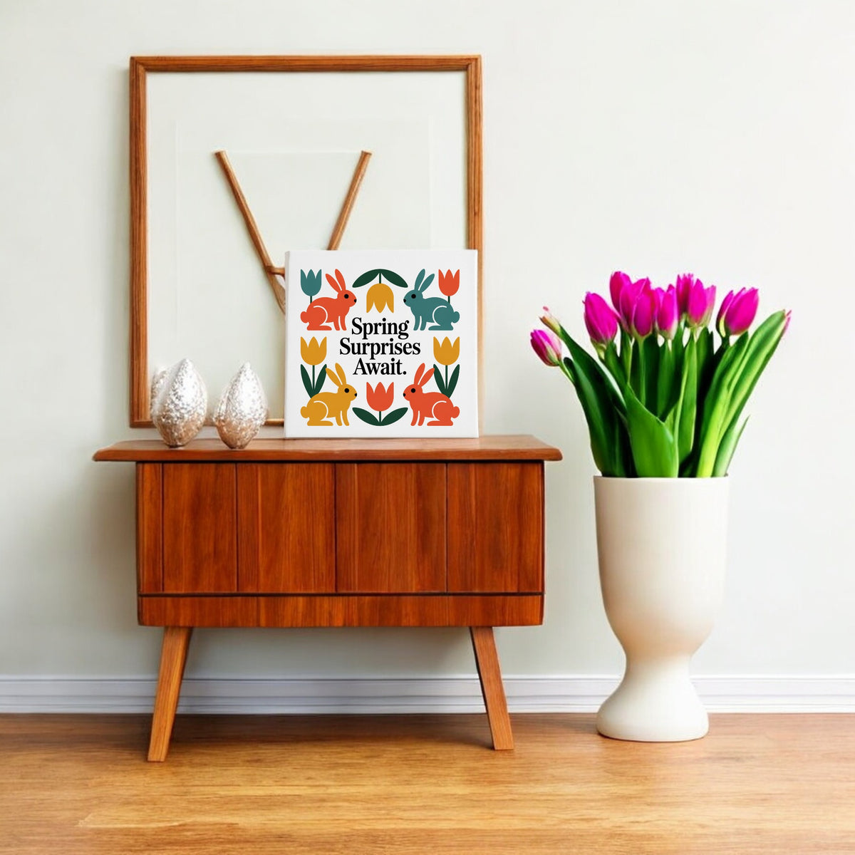 A wooden sideboard with decor sits by a wall. On top are two textured ornaments, Scandinavian-inspired art, and Mirabilia Boutique’s "Spring Surprises Await Canvas – 8x8 Colorful Bunnies." A white vase of pink tulips stands beside the sideboard.