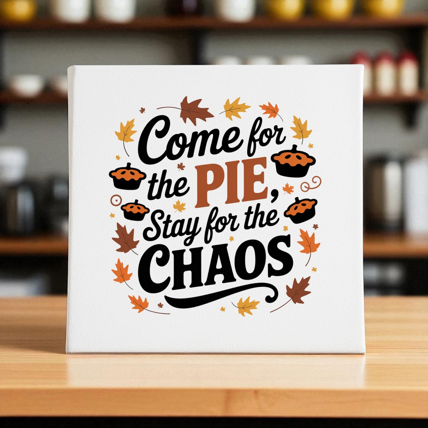 The Mirabilia Boutique "Come for the Pie Chaos Art" canvas features the phrase “Come for the pie, stay for the chaos” with autumn leaves and mini pie illustrations on a wooden counter, set against a blurred kitchen background.