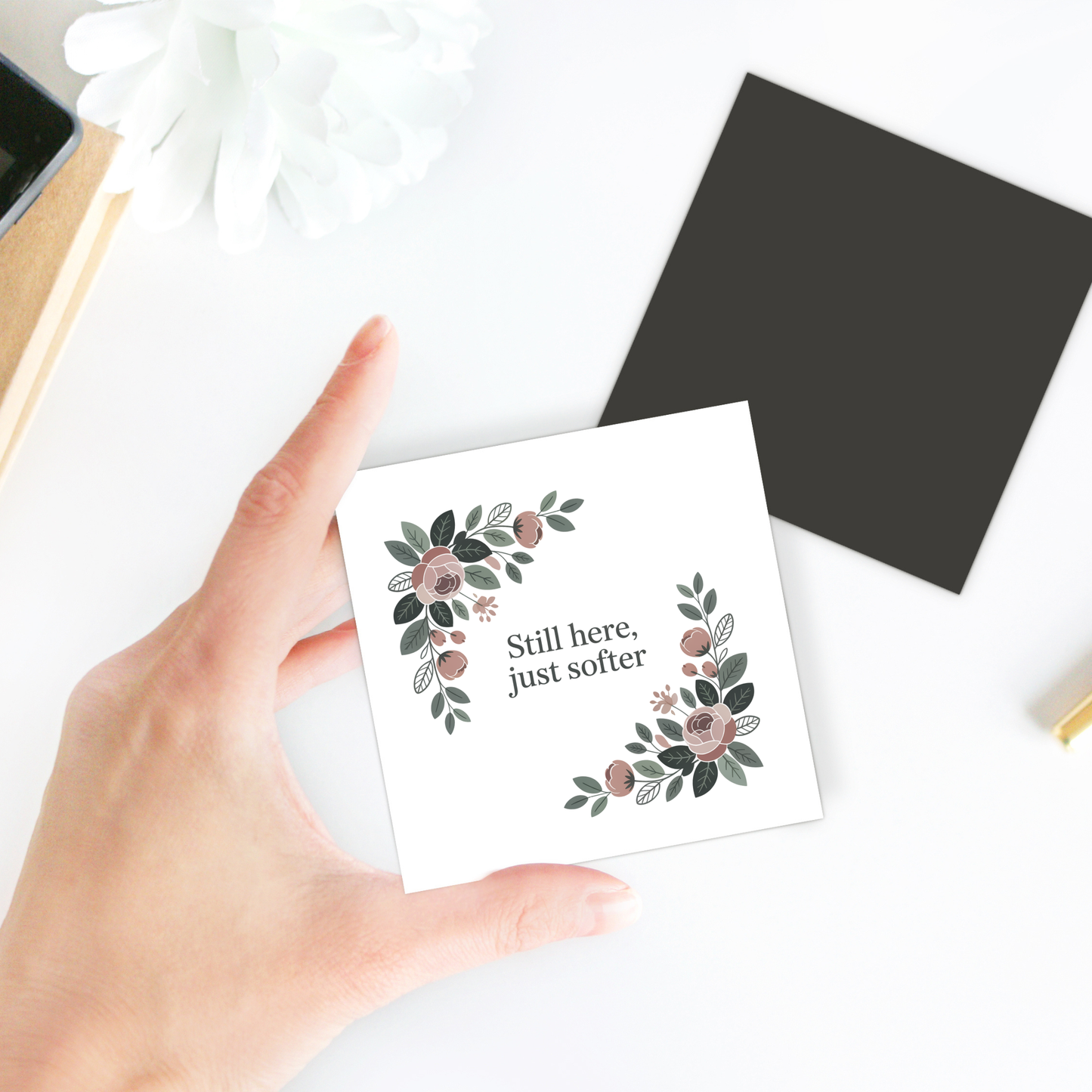 A hand holds the Mirabilia Boutique "Still Here Just Softer Floral Magnet." A black envelope and white flower rest nearby, making this magnet an ideal encouragement gift.