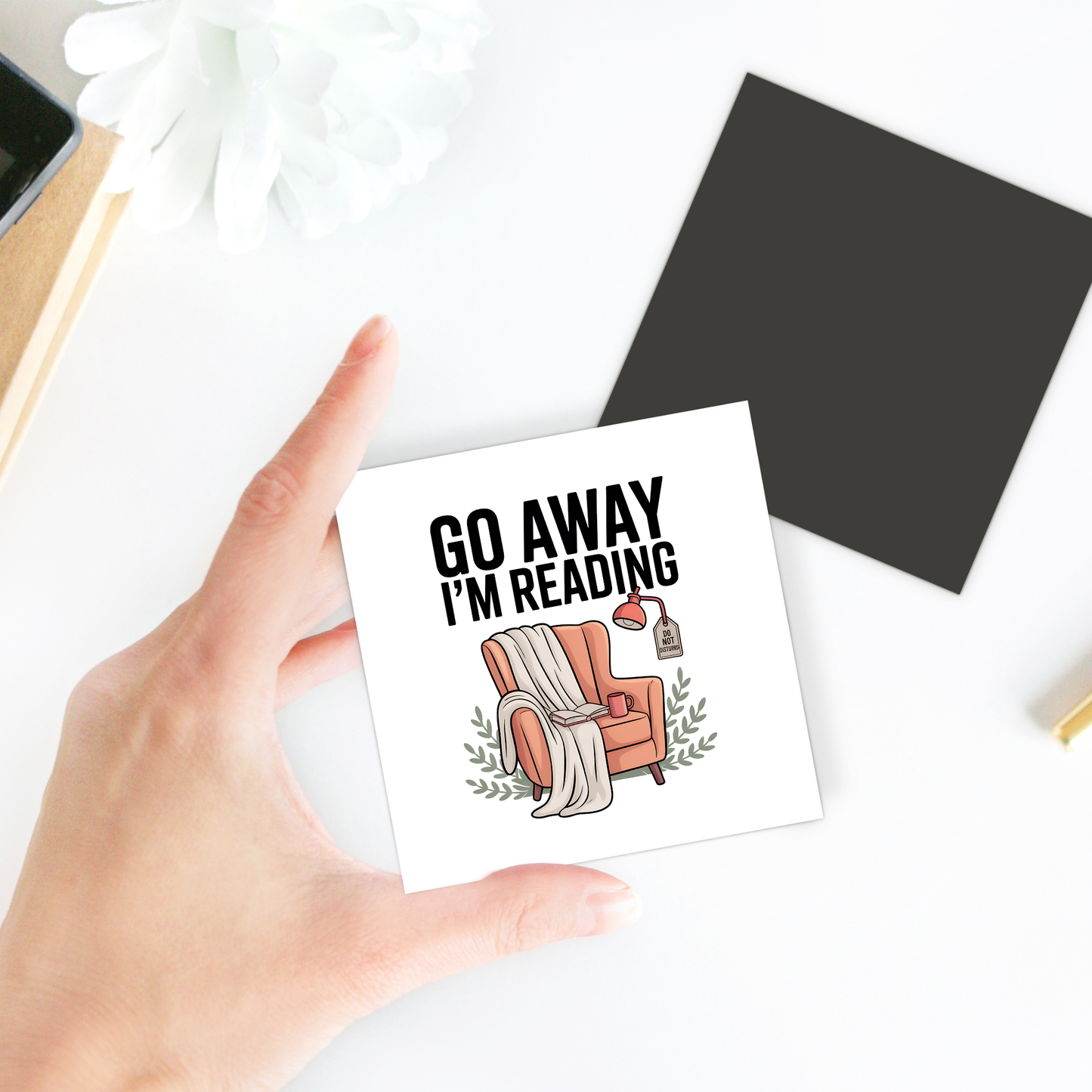 A hand holds the "Go Away I'm Reading Magnet" by Mirabilia Boutique, featuring a cozy armchair, blanket, lamp, and whimsical text—an ideal funny book lover gift or cute bookish decor. A flower, black square, and notebook rest on a white table background.