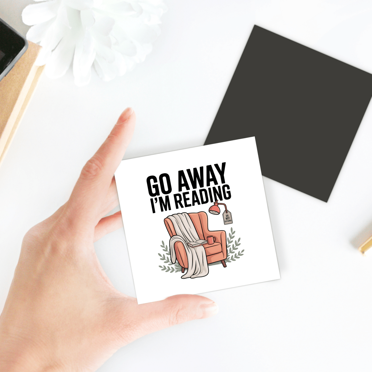 A hand holds the "Go Away I'm Reading Magnet" by Mirabilia Boutique, featuring a cozy armchair, blanket, lamp, and whimsical text—an ideal funny book lover gift or cute bookish decor. A flower, black square, and notebook rest on a white table background.