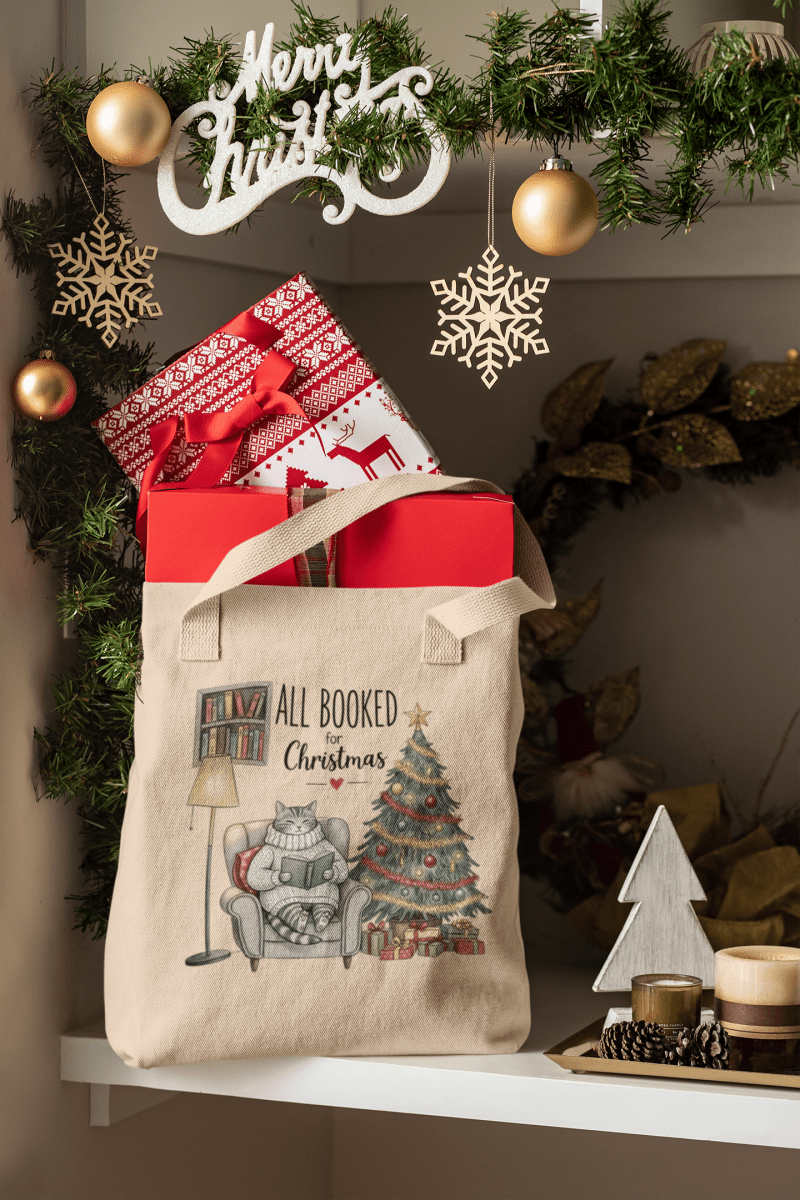 A festive "All Booked for Christmas Cat Tote Bag" by Mirabilia Boutique sits on a shelf with wrapped gifts, golden ornaments, greenery, and wooden holiday decor. The tote features a cat, books, Christmas tree, and presents.