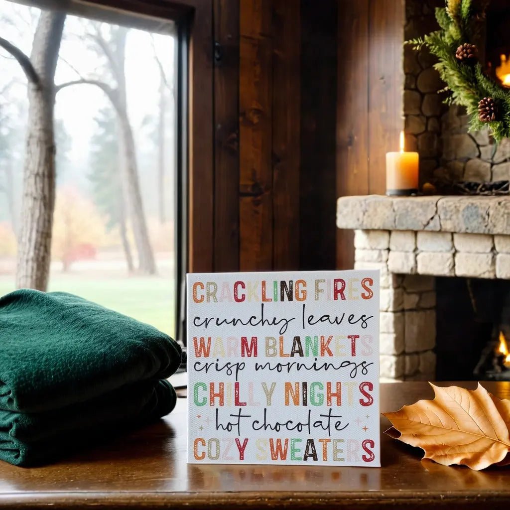 A cozy scene features a green blanket, brown leaf, lit candle by a stone fireplace, and Mirabilia Boutique’s Fall Words Canvas Sign—an 8x8 cradled wood UV printed autumn decor—near a window with foggy trees for perfect fall vibes.