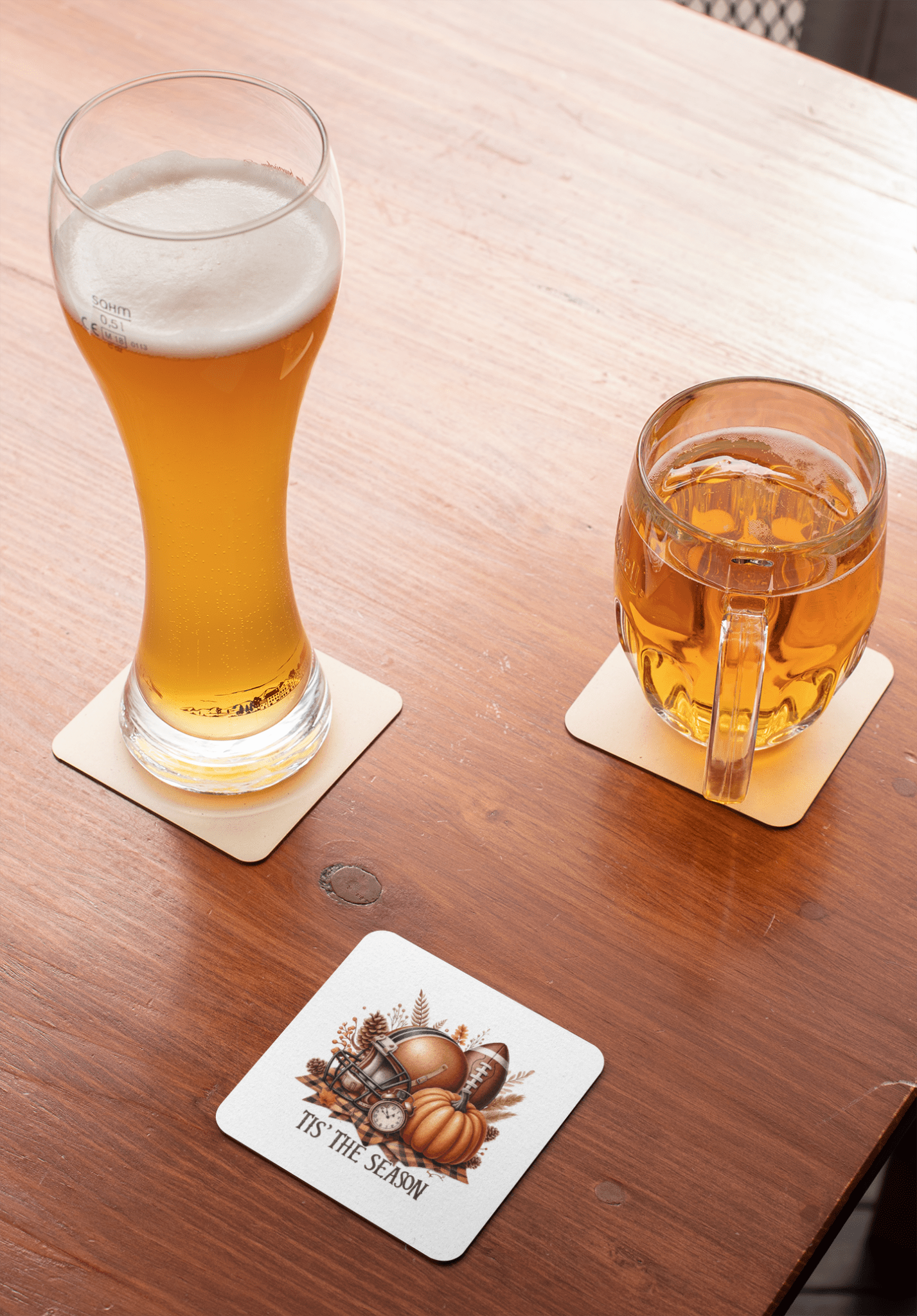 A tall glass of pale beer and a mug of amber beer rest on coasters atop a wooden table. Nearby is a Mirabilia Boutique "Tis’ the Season Coaster" from their Fall Football Pumpkin Drink Decor set.