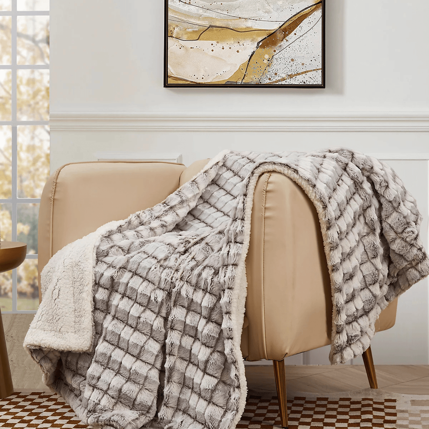 A beige armchair features the Mirabilia Boutique Custom Fall Throw Blanket, plaid-patterned and textured, in a bright room with a large window and abstract art—a perfect cozy autumn gift for loved ones.