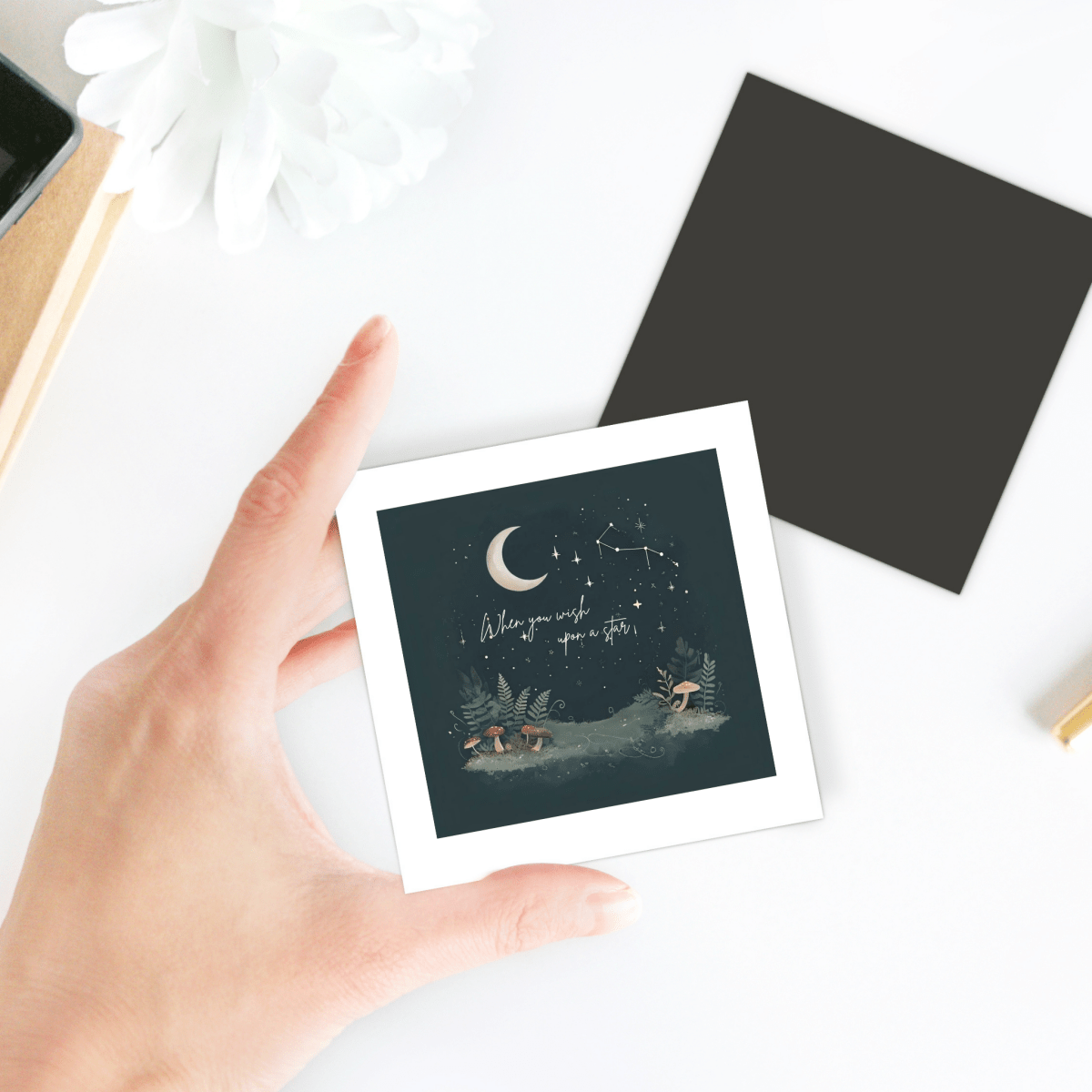 A hand holds the "When You Wish Upon a Star Woodland Magnet" by Mirabilia Boutique—a square magnet showcasing a night sky with a crescent moon, stars, constellations, and the phrase "Make your wish upon a star" above a peaceful forest scene. This enchanting piece of celestial decor is perfect for any cottagecore gift collection.