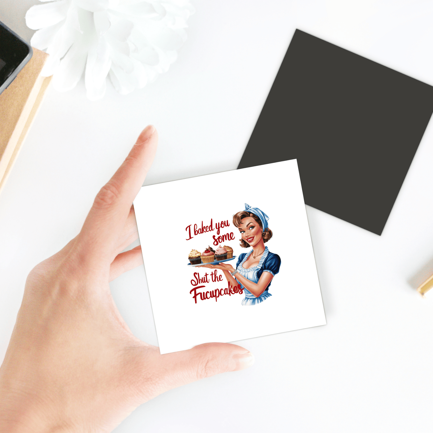 A hand holds the Mirabilia Boutique Fucupcakes Magnet, a funny retro 3x3" UV printed kitchen fridge magnet featuring a woman with cupcakes and the caption "I baked you some Shut the Fucupcakes." A black square and white flower are also nearby.
