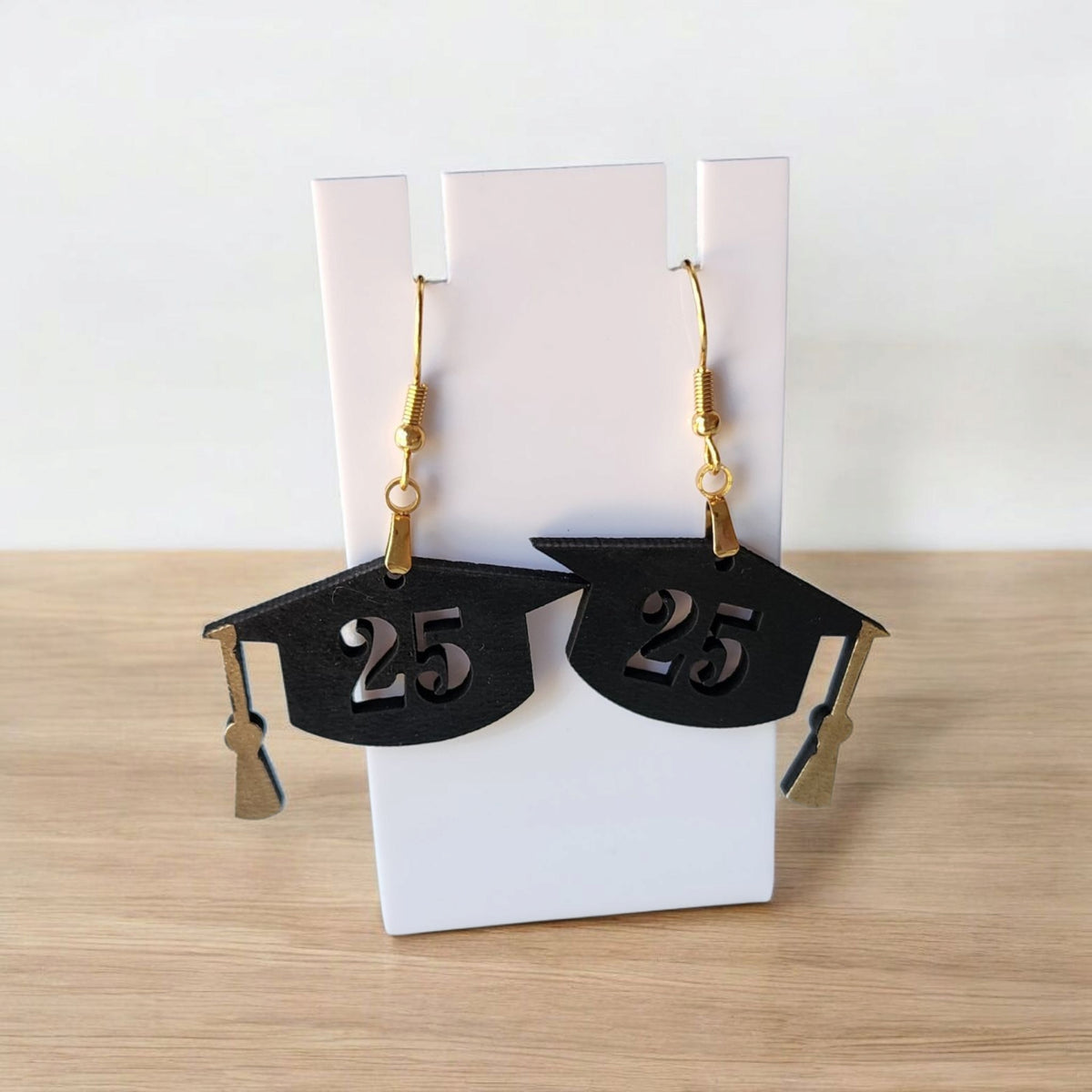 Mirabilia Boutique's Graduation Cap Earrings Class of 2025 feature black laser-cut caps with "25," gold hooks, and tassels—perfect grad gifts or party accessories—shown on a white stand against a light wood surface.