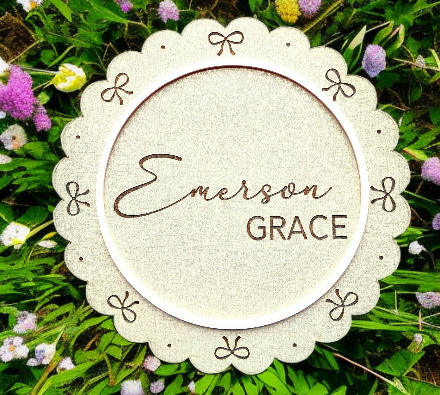 The Personalized Baby Name Shelf Sign by Mirabilia Boutique features a round, scalloped edge with bows and “Emerson Grace” in script—perfect nursery shelf decor set against green leaves and pink, white, and purple flowers.