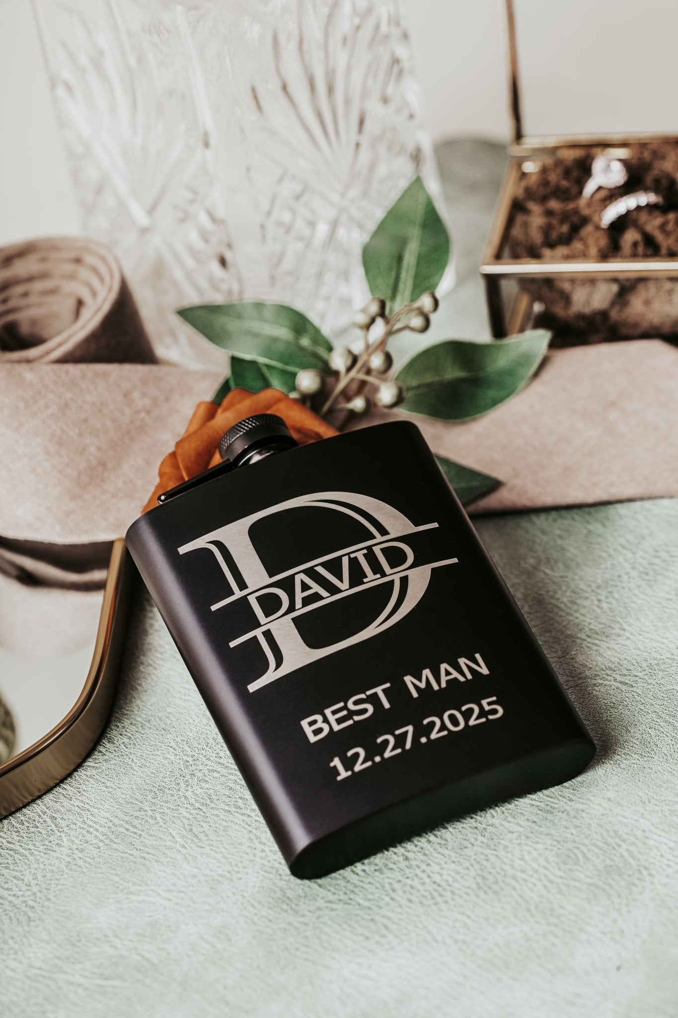 A Mirabilia Boutique Personalized Best Man Flask Gift Set engraved with "David, Best Man, 12.27.2025" sits on green fabric among a tan belt and glass container—an ideal custom groomsmen present and keepsake.