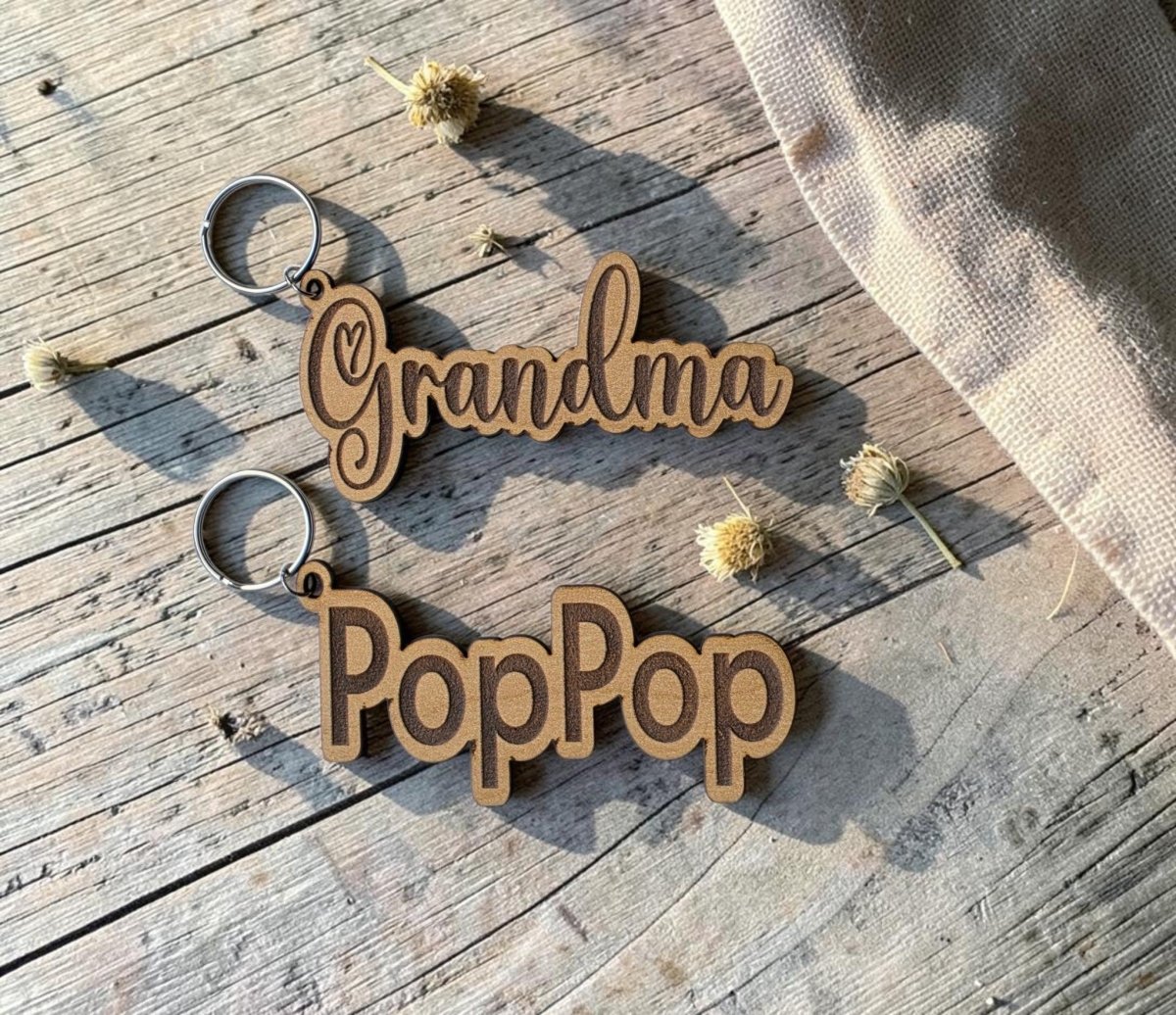 Two Personalized Engraved Wood Name Keychains from Mirabilia Boutique rest on a wooden surface—one featuring “Grandma” elegantly engraved in cursive, the other boldly displaying “PopPop”—with dried flowers and beige fabric accents nearby.