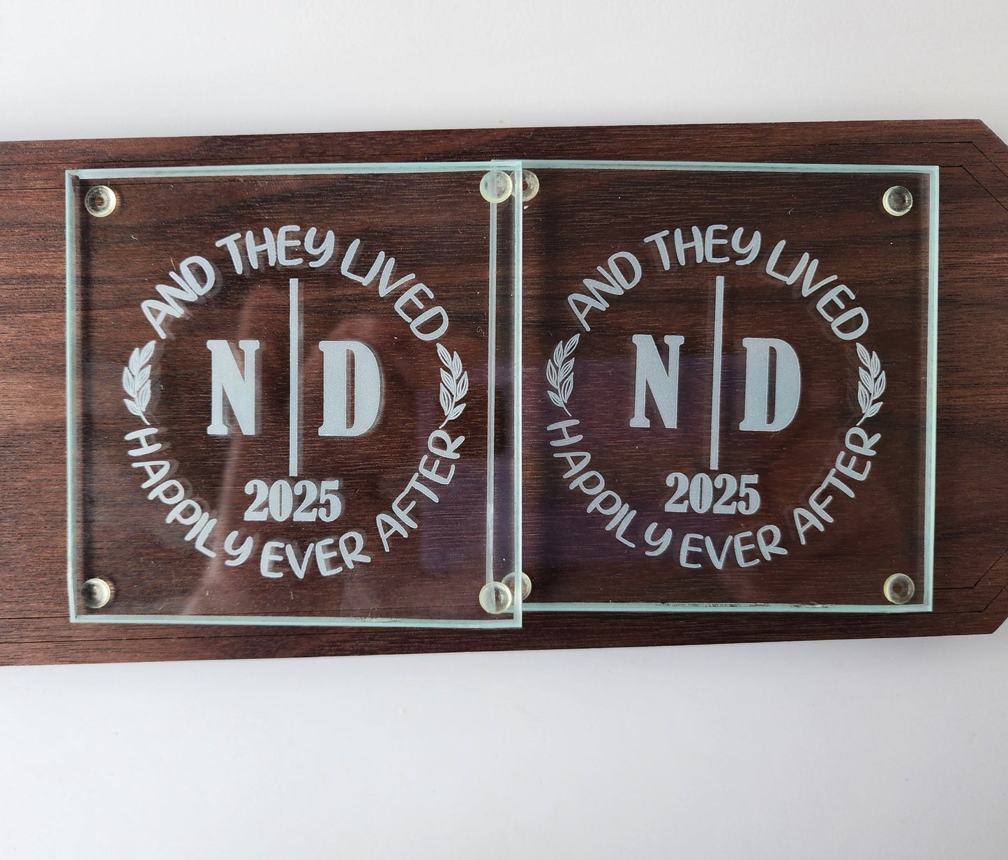 The Personalized Glass Wedding Coasters by Mirabilia Boutique elegantly display "And They Lived Happily Ever After," featuring the initials "ND" and the year "2025," making a unique and perfect personalized wedding gift.