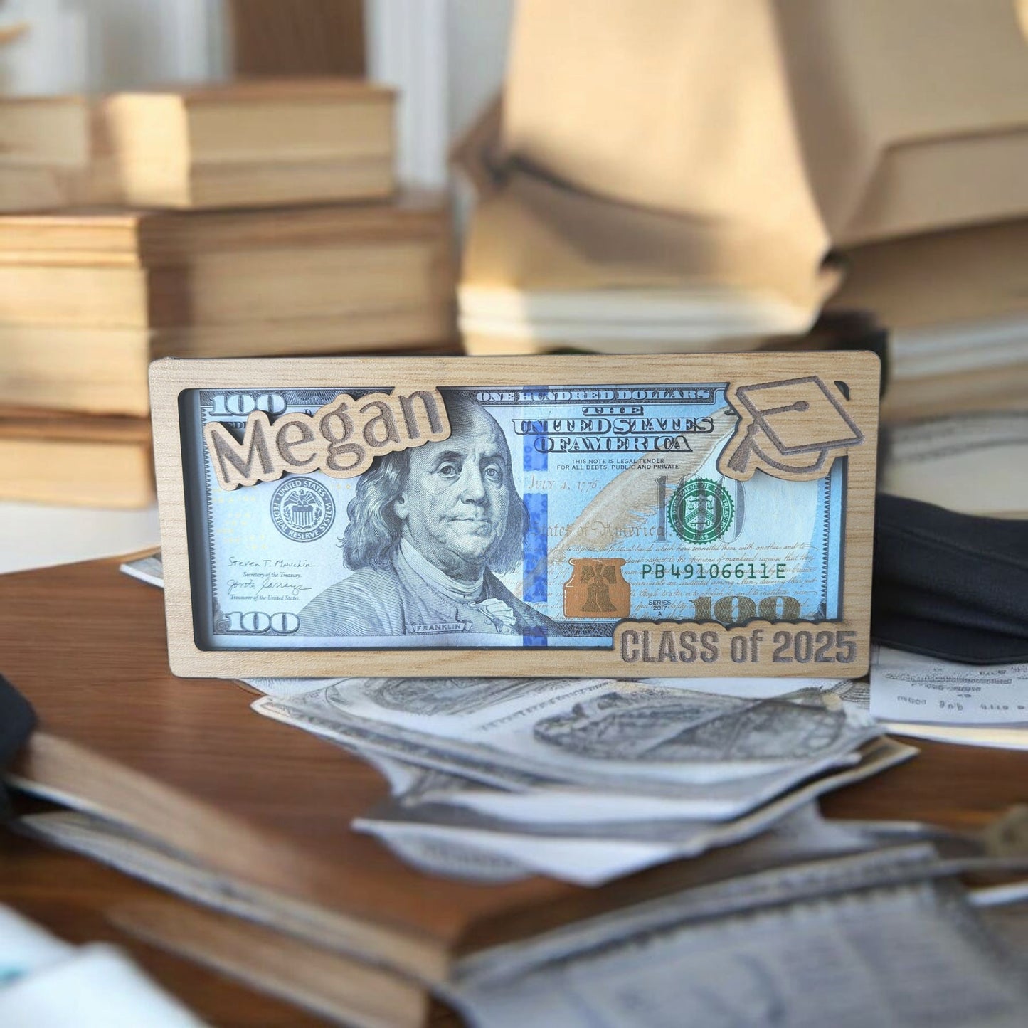 The Mirabilia Boutique Personalized Graduation Money Holder is a custom wood cash gift for the Class of 2025, featuring decorative "Megan" and a graduation cap—an ideal, unique present displayed on a desk amidst books and papers.