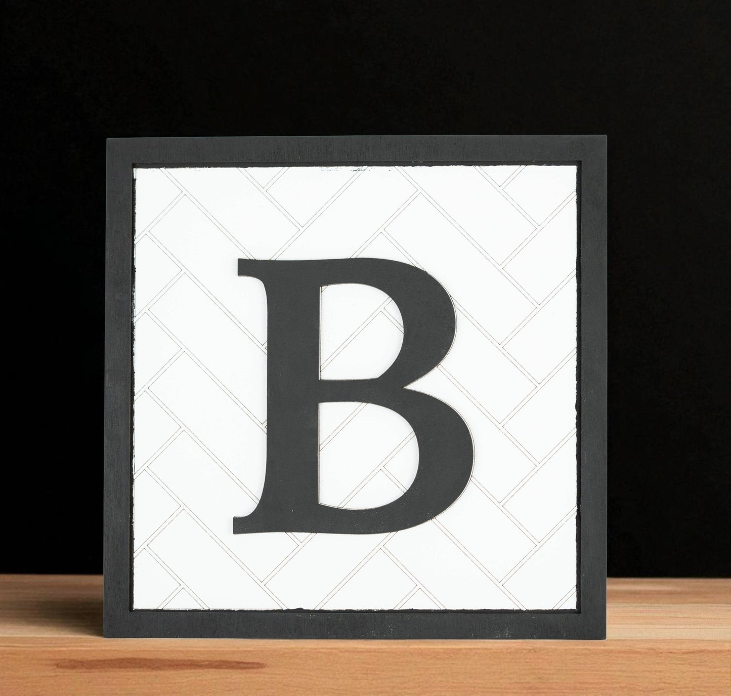 The Mirabilia Boutique Custom Initial Shelf Sign | Herringbone Backer | 10x10 displays a bold black letter "B" on a white herringbone background—ideal personalized decor for any modern farmhouse, shown on light wood against a black backdrop.