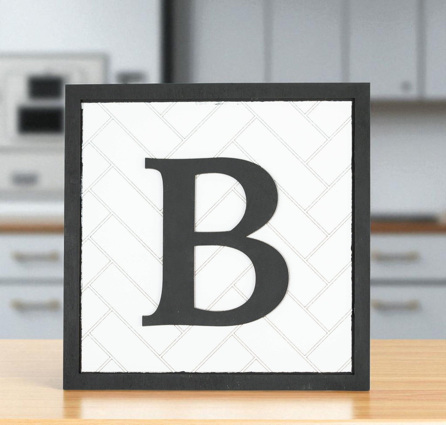 The Mirabilia Boutique Custom Initial Shelf Sign | Herringbone Backer | 10x10 features a bold black “B” on a white background with a black frame, adding personalized decor to any modern kitchen countertop.