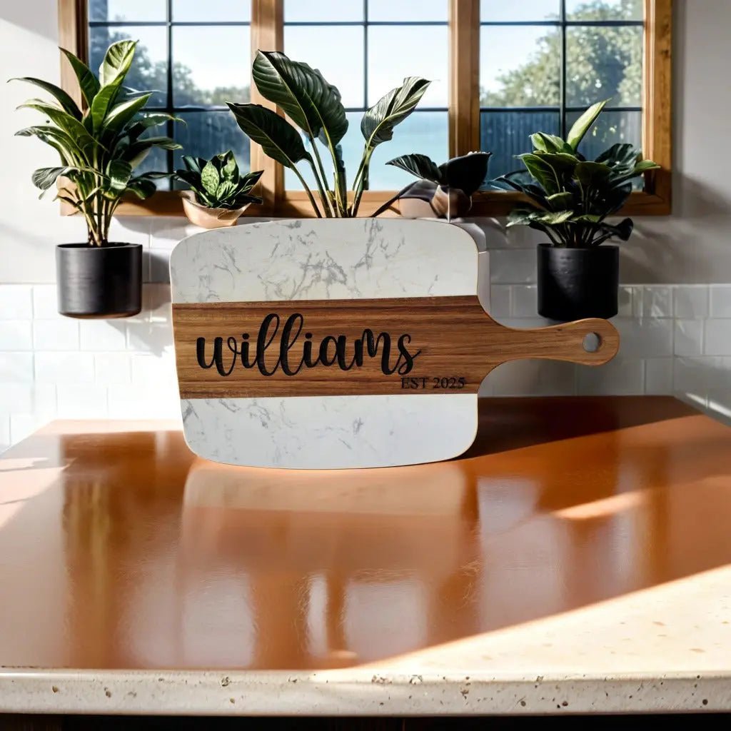 The Mirabilia Boutique Personalized Marble and Acacia Serving Board, engraved with "Williams" and "EST 2023," sits on a shiny brown counter as a stylish charcuterie centerpiece. Three potted plants decorate the windowsill in the background.