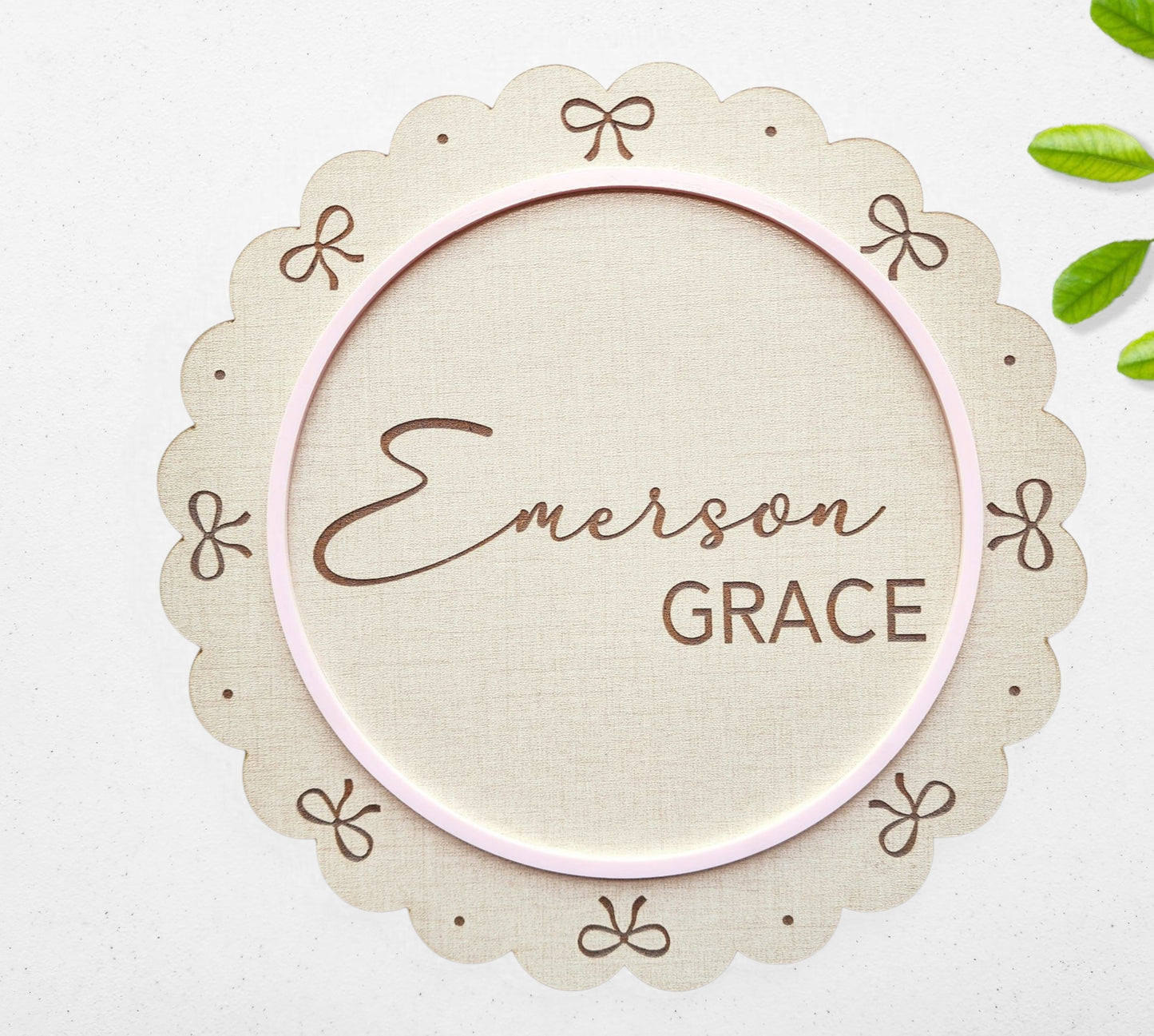 The Mirabilia Boutique Personalized Baby Name Shelf Sign features scalloped edges, bow accents, and a pink oval border. Custom with your baby's name, this beige nursery decor includes green leaf details—perfect for gifting or displaying on a shelf.
