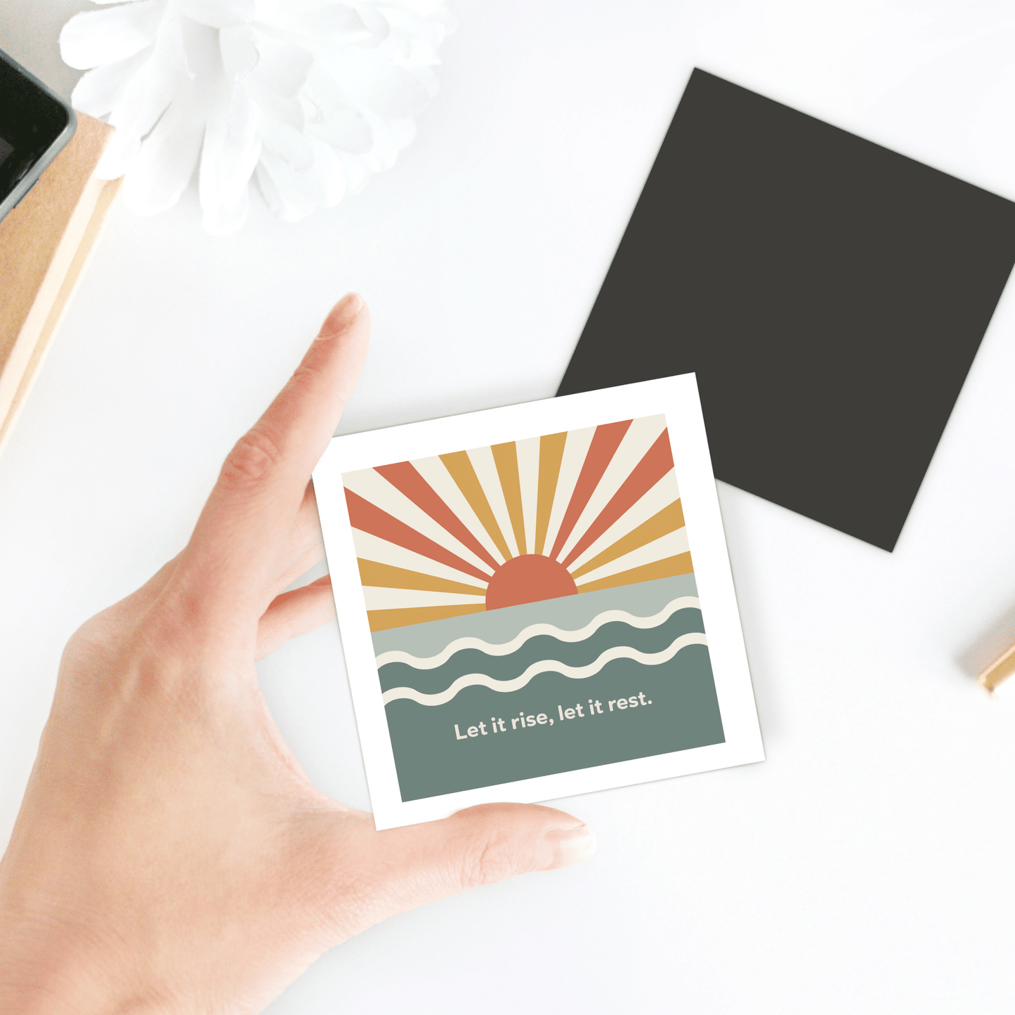 A hand holds the Mirabilia Boutique Let It Rise, Let It Rest Magnet – Sunrise Affirmation, a square magnet with a sunrise over waves and text “Let it rise, let it rest.” Nearby are other objects and a black square on the white surface.