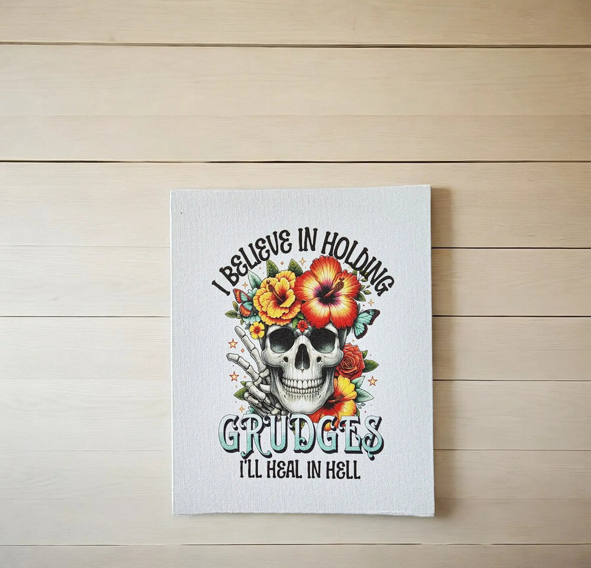 The Mirabilia Boutique "I Believe in Holding Grudges" 8x10 canvas print features a skull with colorful flowers and bold text on a light wooden background—dark humor decor for making a statement in any space.
