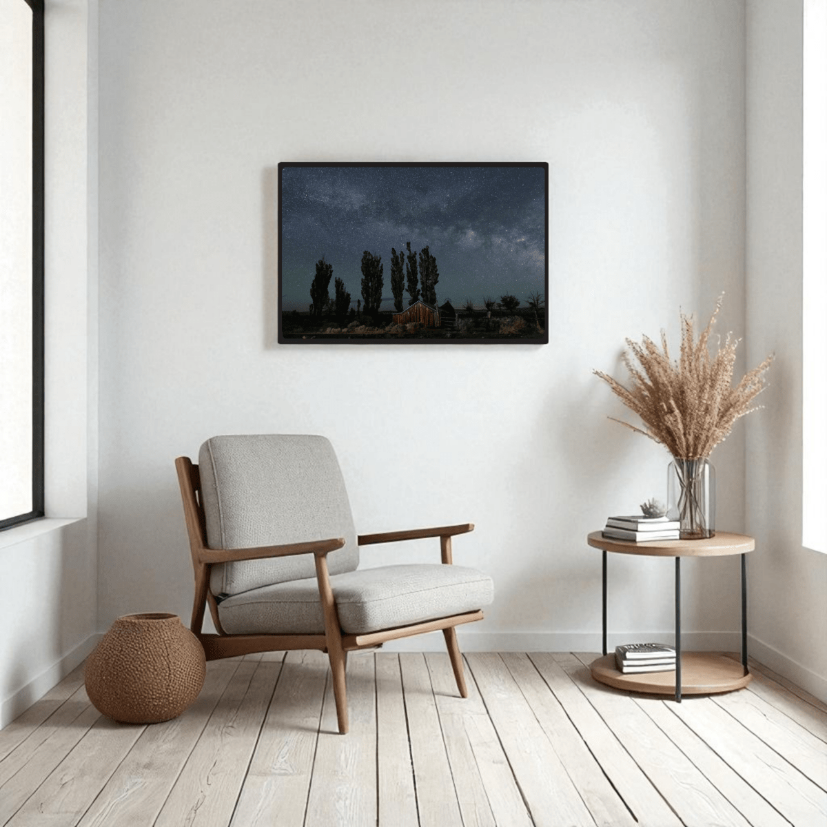 A modern, minimalist living room with a gray armchair, round side table adorned with dried grass, woven basket, and the DeChambeau Ranch Milky Way Print by Mirabilia Boutique—an archival-quality framed fine art print of the Eastern Sierra night sky—hanging on the wall above the chair.