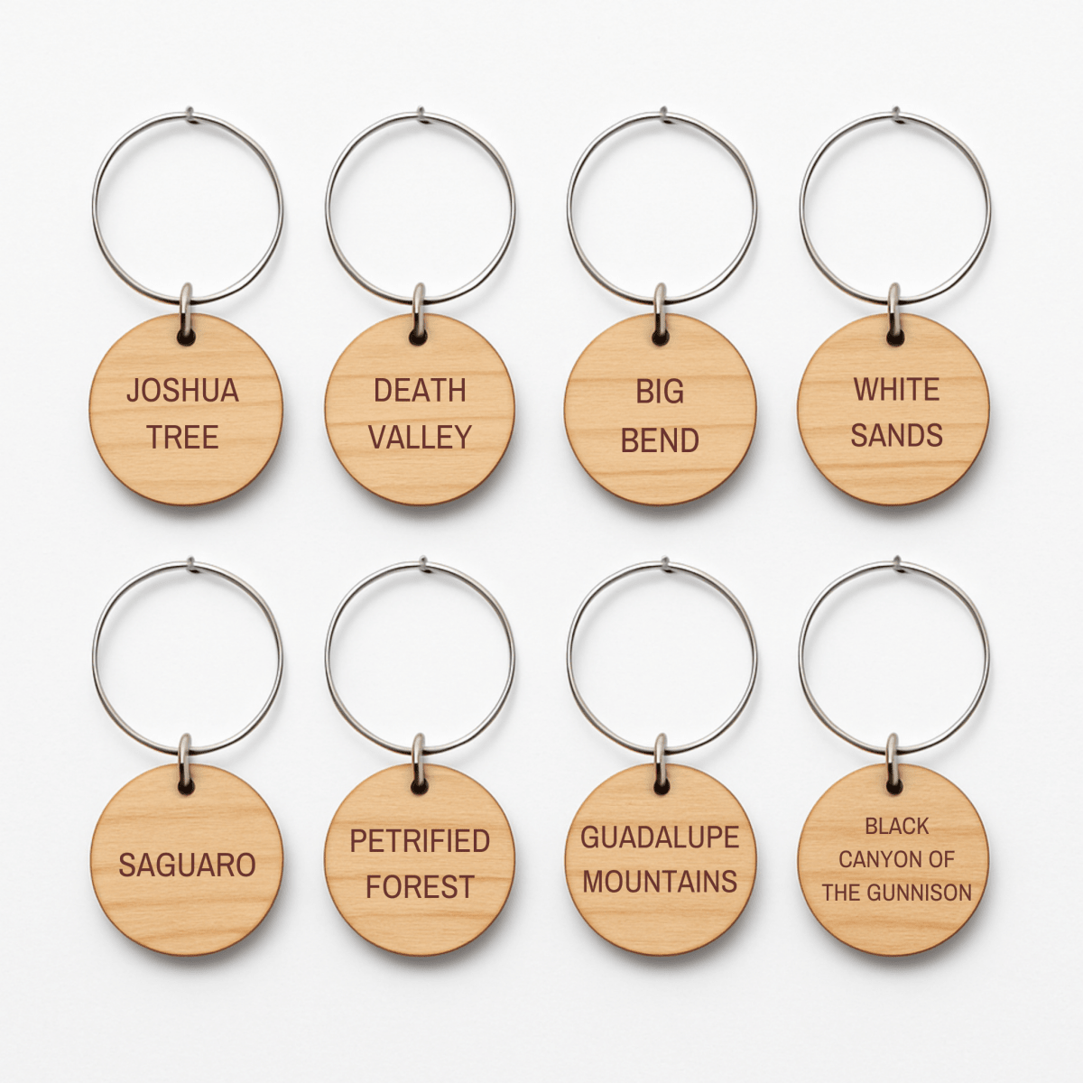 Eight round wooden wine charms from Mirabilia Boutique’s Desert Dreaming Wine Charms collection are arranged in two rows, each engraved with a different U.S. national park name like Joshua Tree, Death Valley, and White Sands.